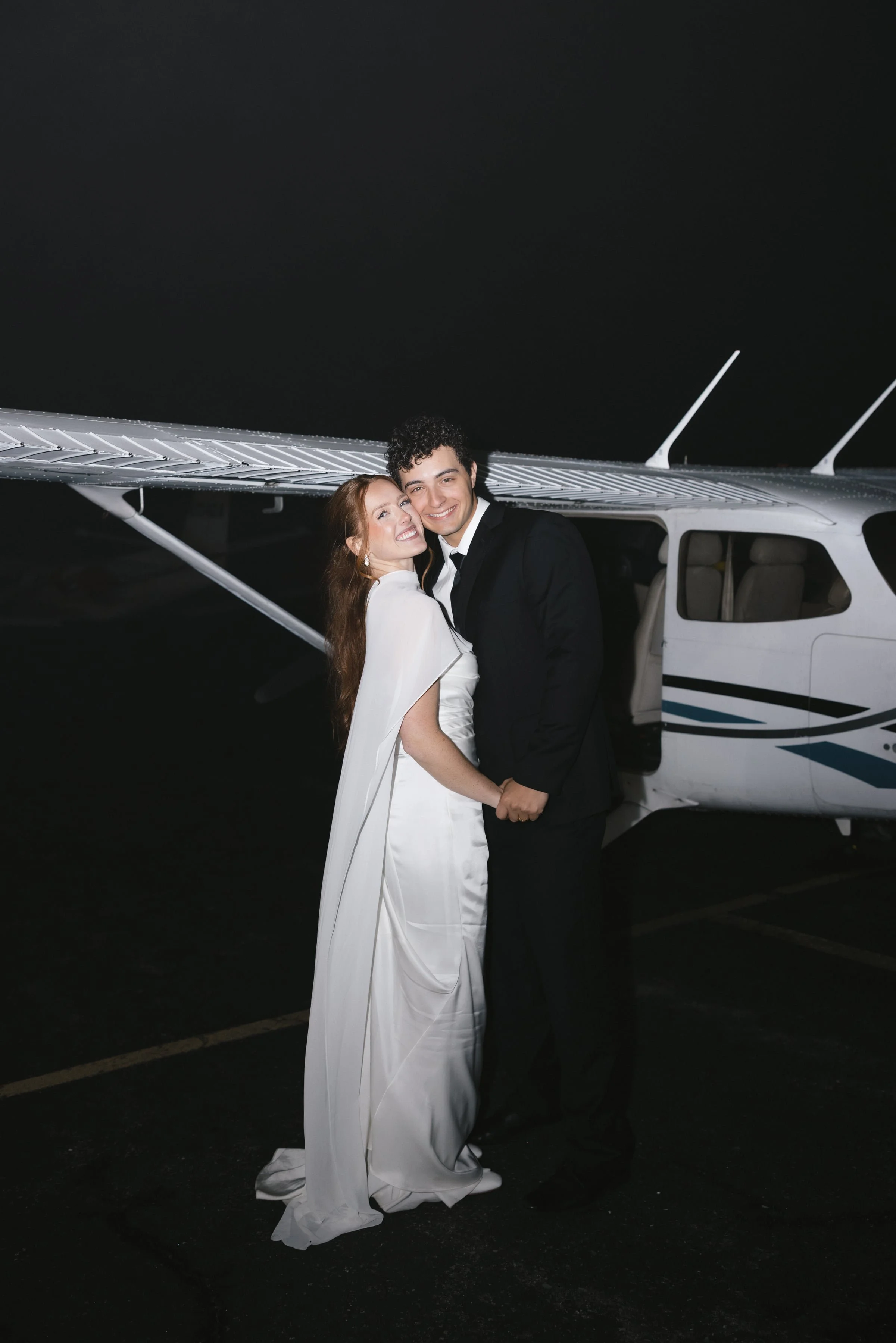 A smiling bride and groom holding hands in front of a small airplane at night, with a dark sky above.
