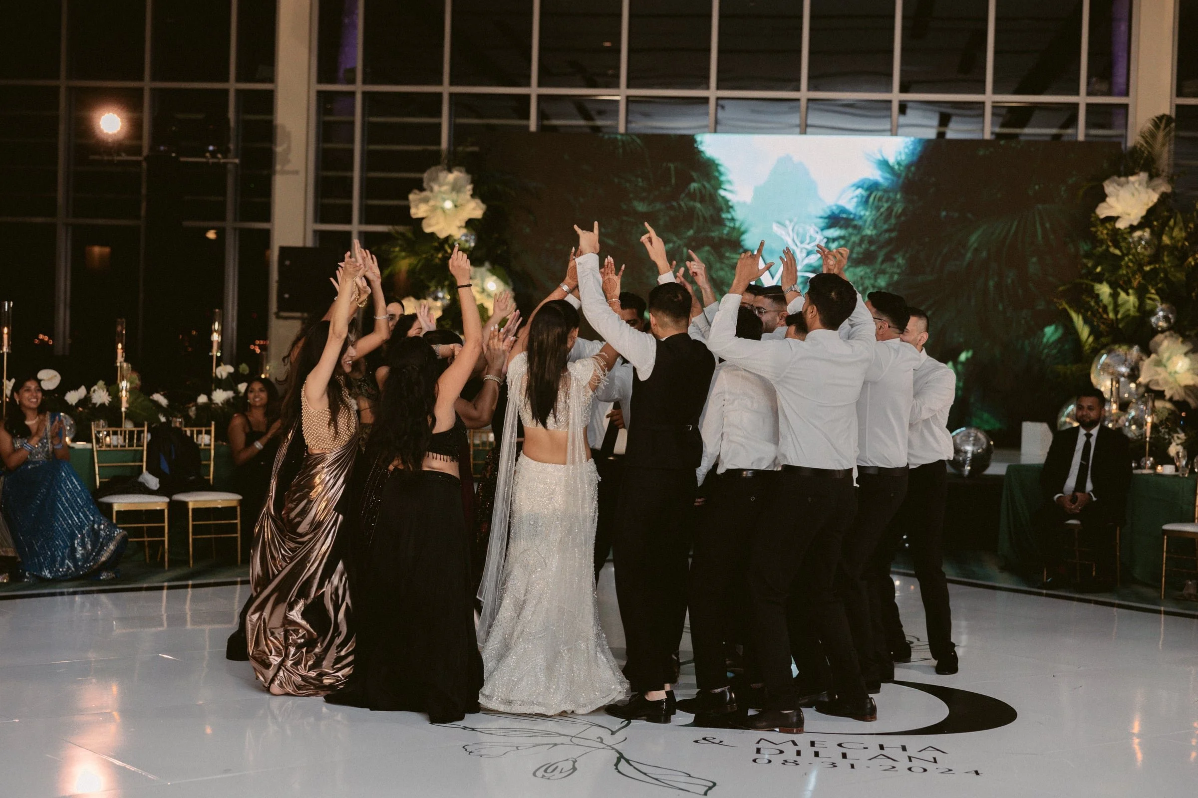 A group of people dancing at a wedding reception, with a bride and groom in the center, a large digital screen with a jungle background behind them, and romantic floral decorations.