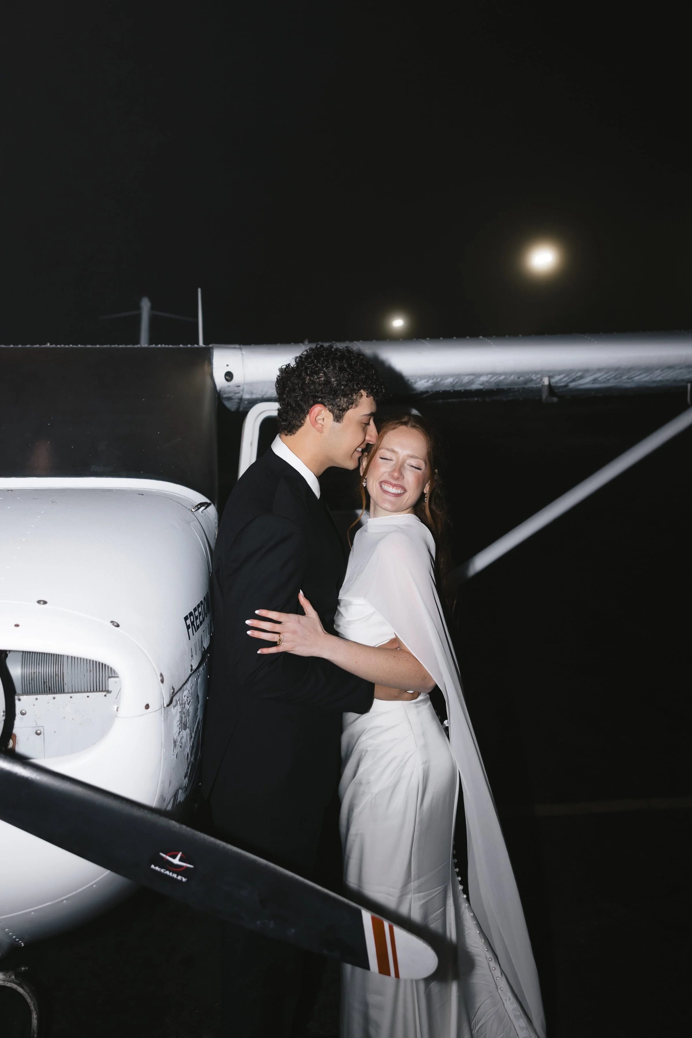 A couple dressed in formal wedding attire embracing next to a small aircraft at night, with the moon visible in the sky.