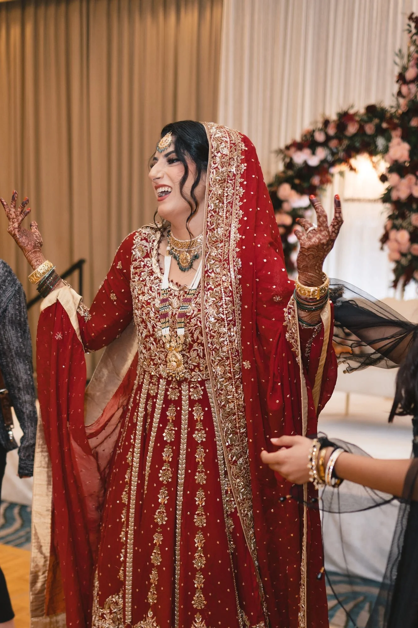 A woman dressed in traditional red and gold Indian bridal attire, smiling with her hands raised, surrounded by floral decorations.