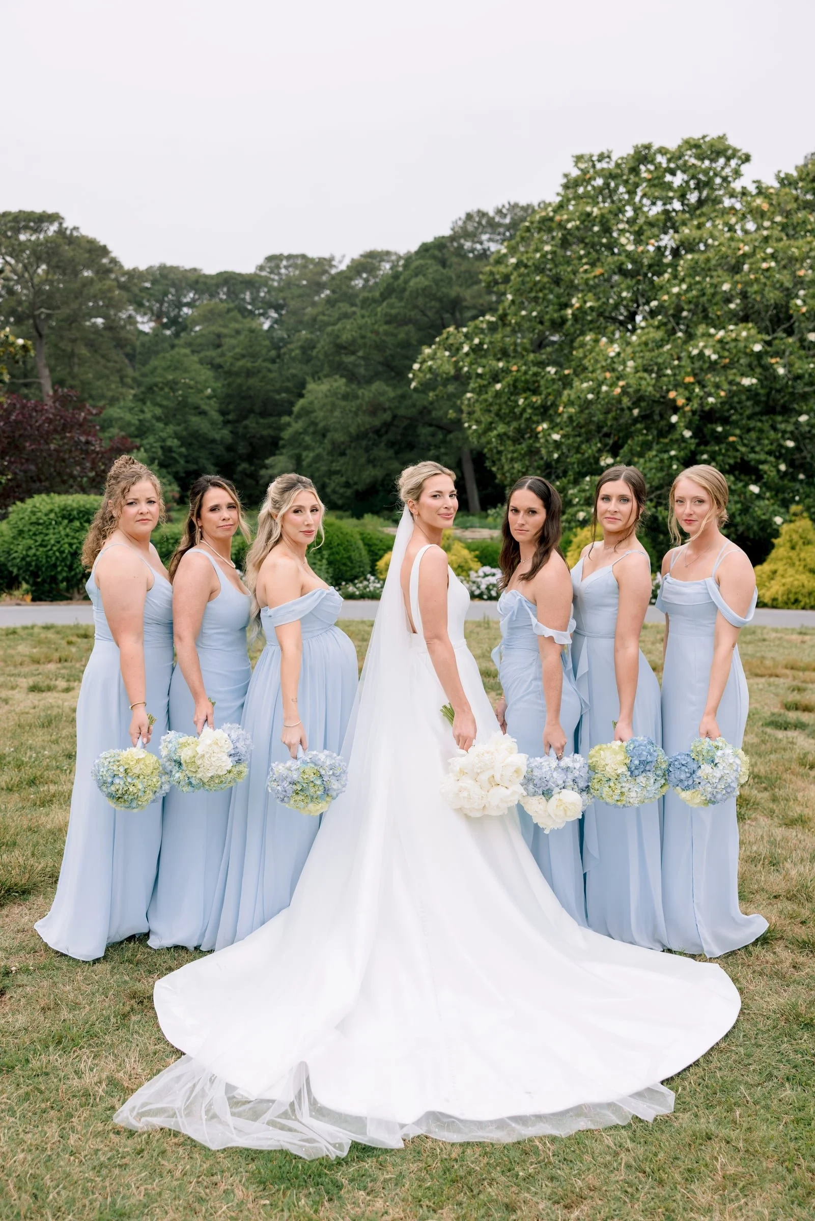 A bride and six bridesmaids standing outdoors on a grassy area with trees and bushes in the background. The women are holding bouquets of white and blue flowers, wearing matching light blue dresses, and looking at the camera.