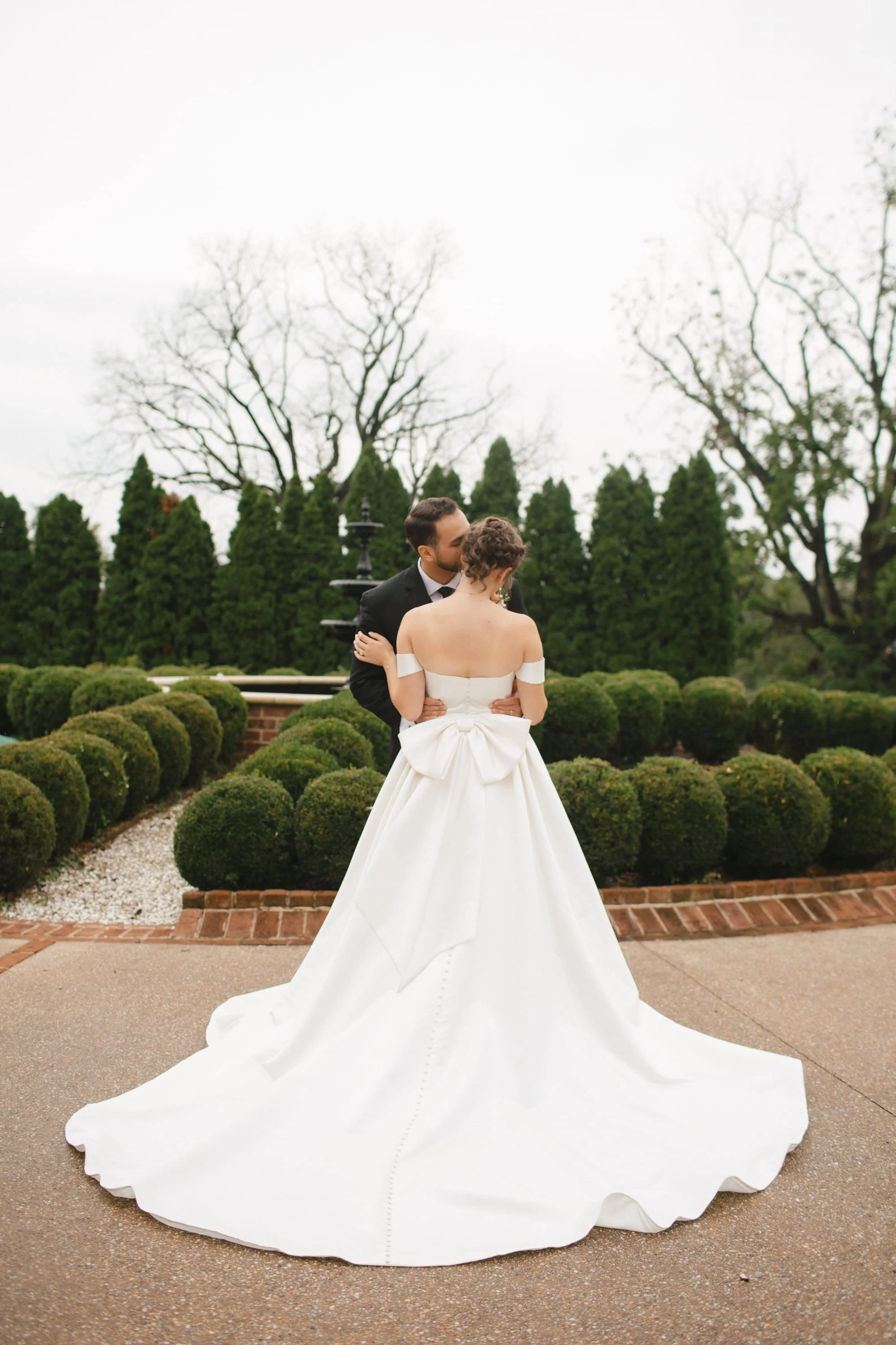 A couple in wedding attire standing outdoors in front of a hedge and trees, sharing a kiss. The woman is in a white off-the-shoulder wedding gown with a large bow at her back and a long train. The man is in a black suit and tie.