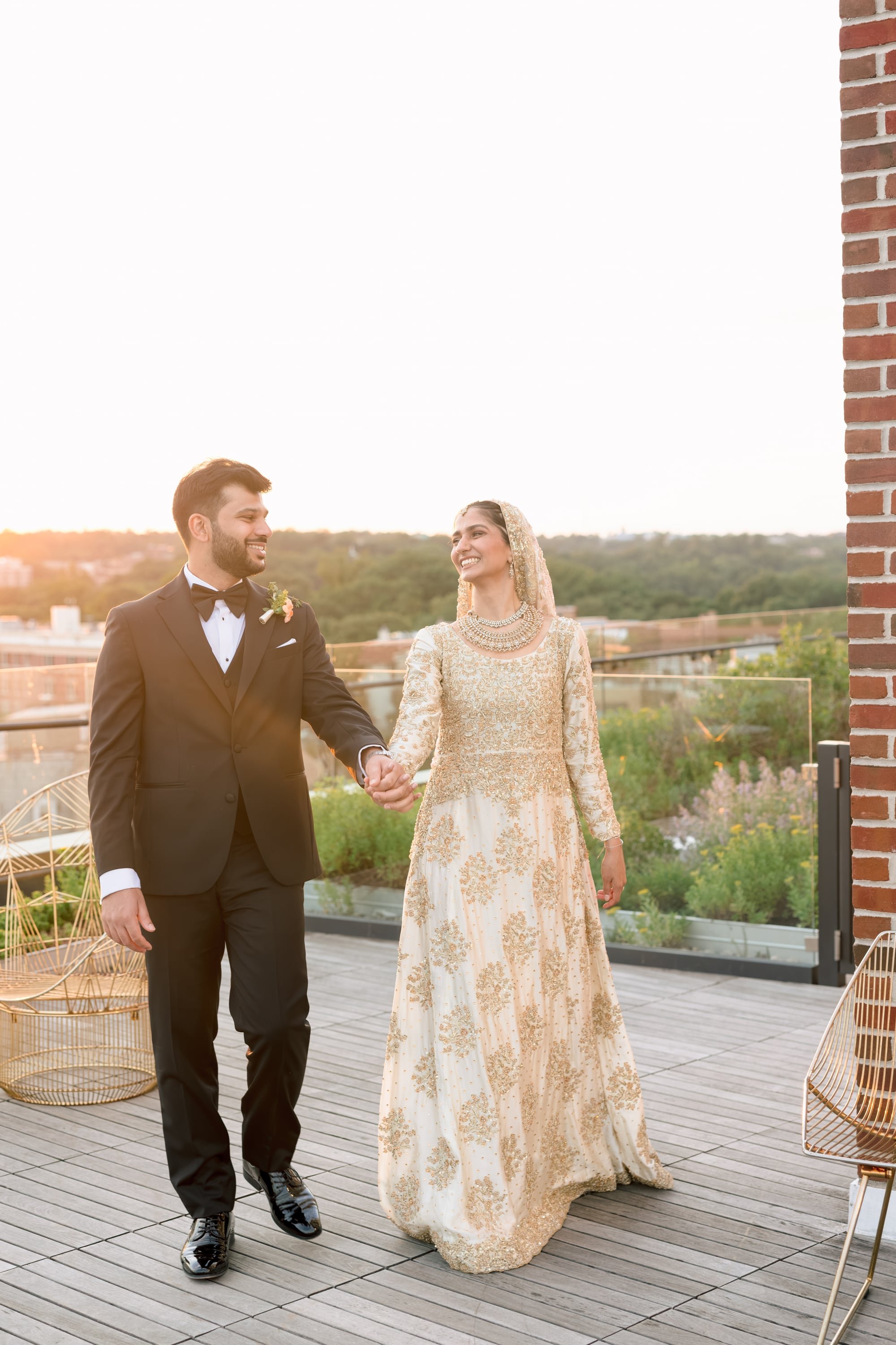 A bride and groom holding hands on a rooftop during sunset. The groom is wearing a black tuxedo with a bow tie, and the bride is dressed in a gold-embellished gown with traditional jewelry and a veil. They are smiling and looking at each other with a