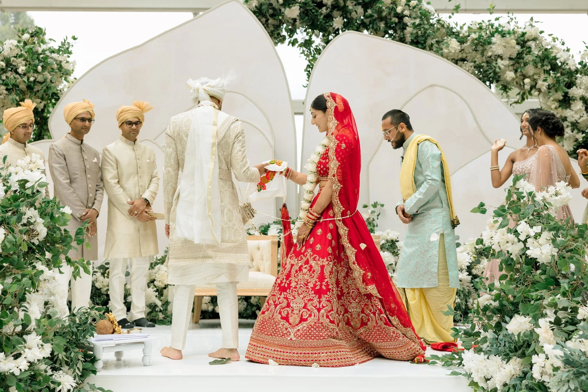 A traditional Indian wedding ceremony taking place outdoors with the bride in a red saree and the groom in cream-colored attire. They are exchanging vows, surrounded by floral decorations and guests dressed in traditional Indian clothing, with a back