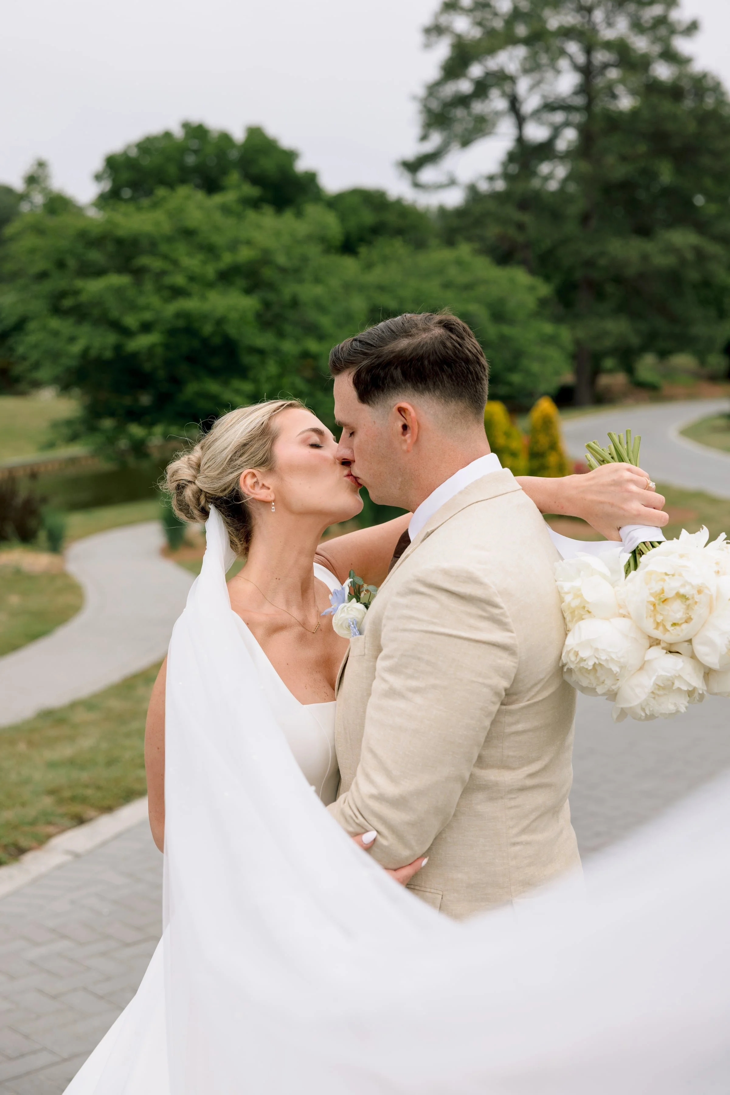 A newlywed couple sharing a kiss outdoors during daytime, with the bride in a white wedding gown and the groom in a beige suit, holding a bouquet of white flowers, greenery, and a floral arrangement. They are on a paved pathway with lush green trees 