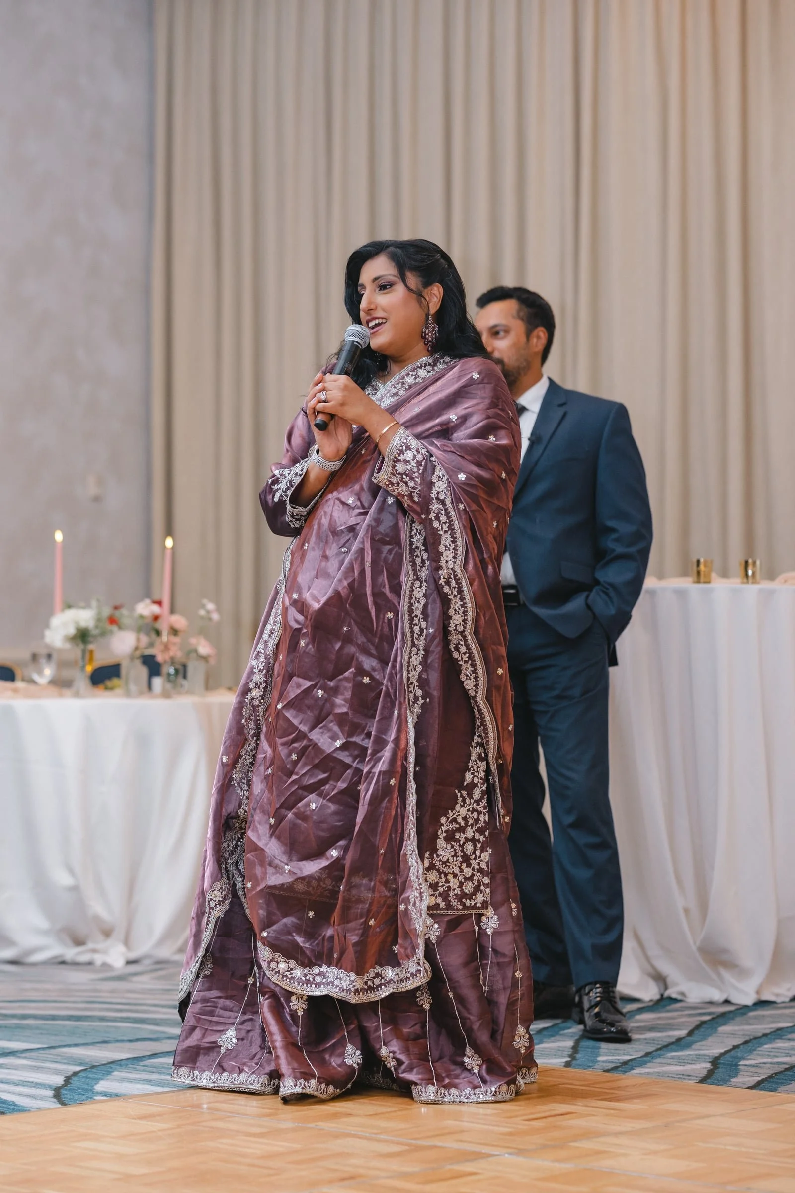 A woman in a traditional purple saree holding a microphone, speaking at an event with a man in a suit standing behind her. The background features beige curtains and decorated tables with candles and flowers.