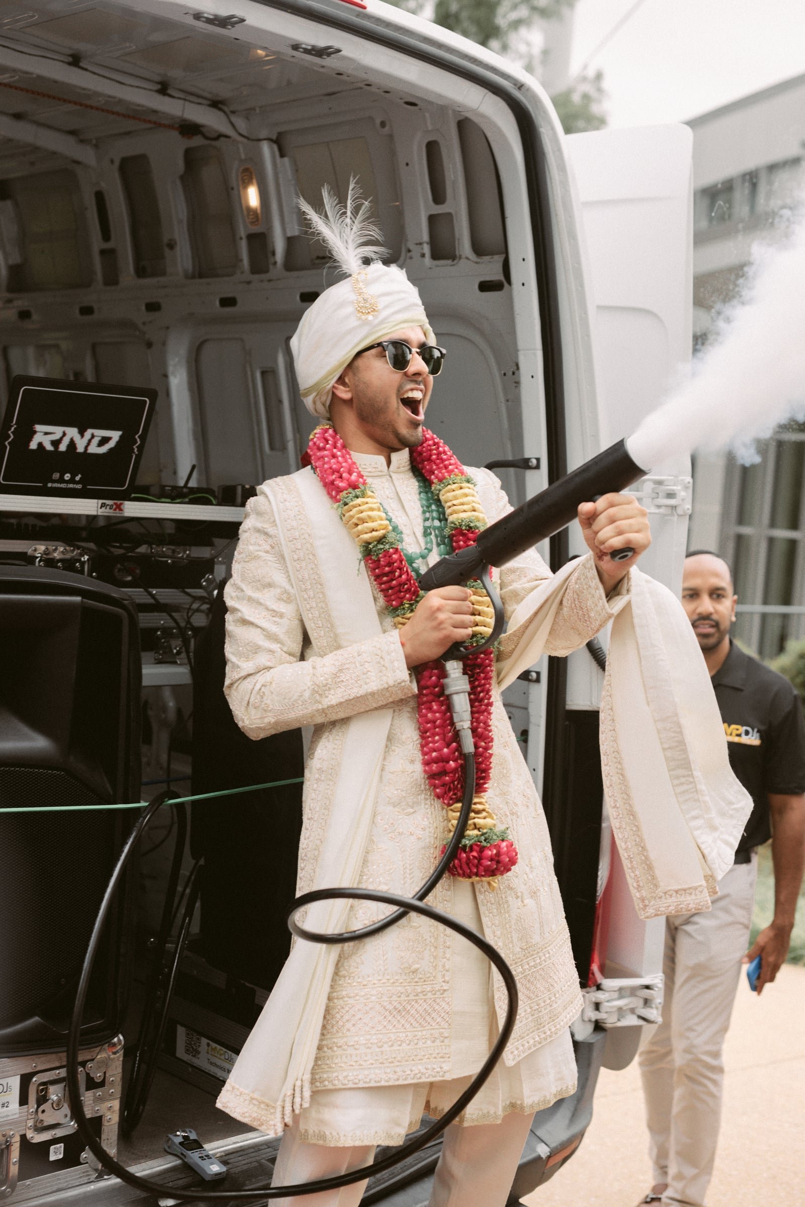 A person dressed in traditional Indian attire, wearing sunglasses and a turban with a feather, holding a party cannon that is releasing confetti or smoke, standing in front of a van with DJ equipment, with a security guard and another person visible 