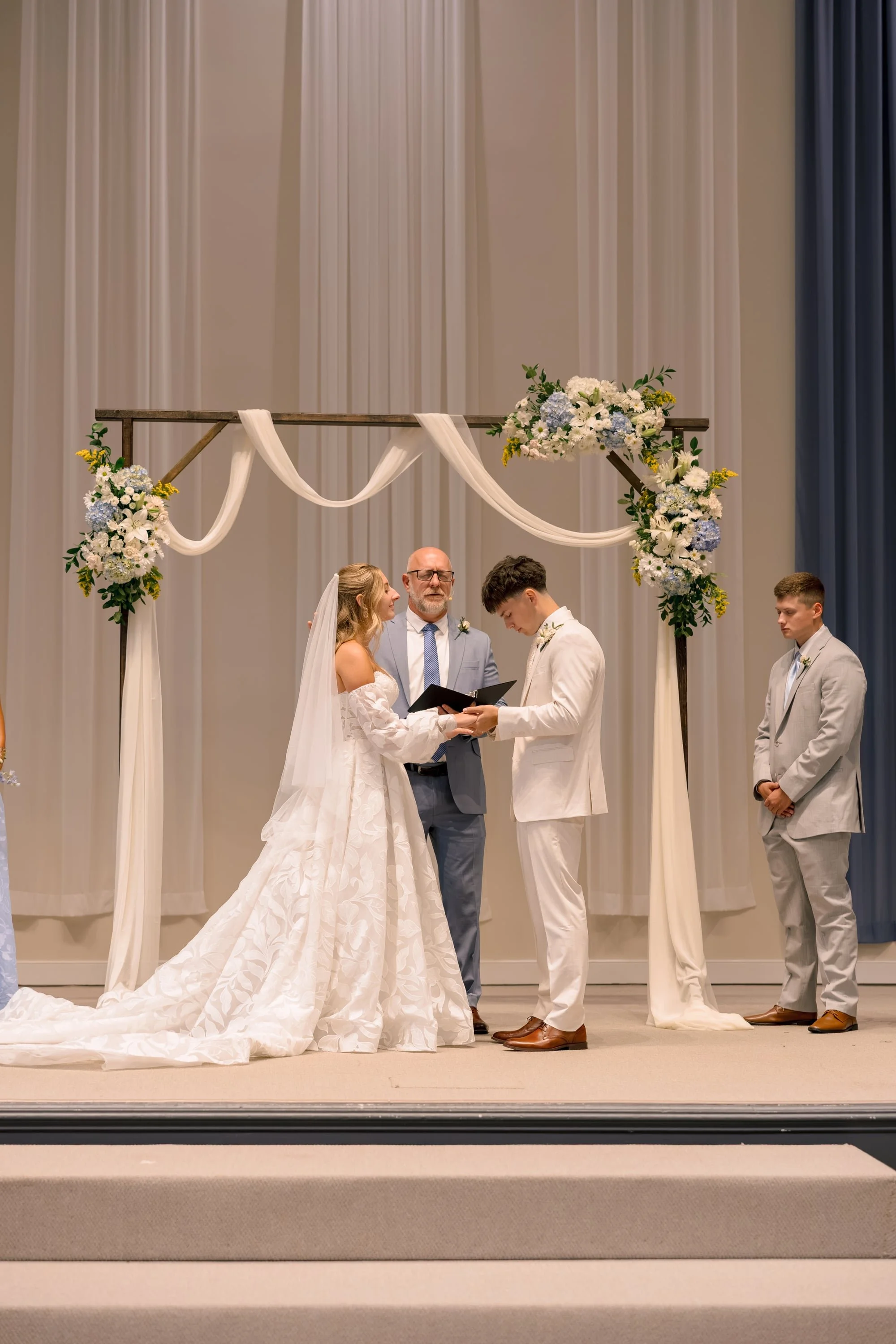 Bride and groom exchanging vows during their wedding ceremony, standing in front of an officiant, with two groomsmen on the side, under a floral arch with draped fabric.