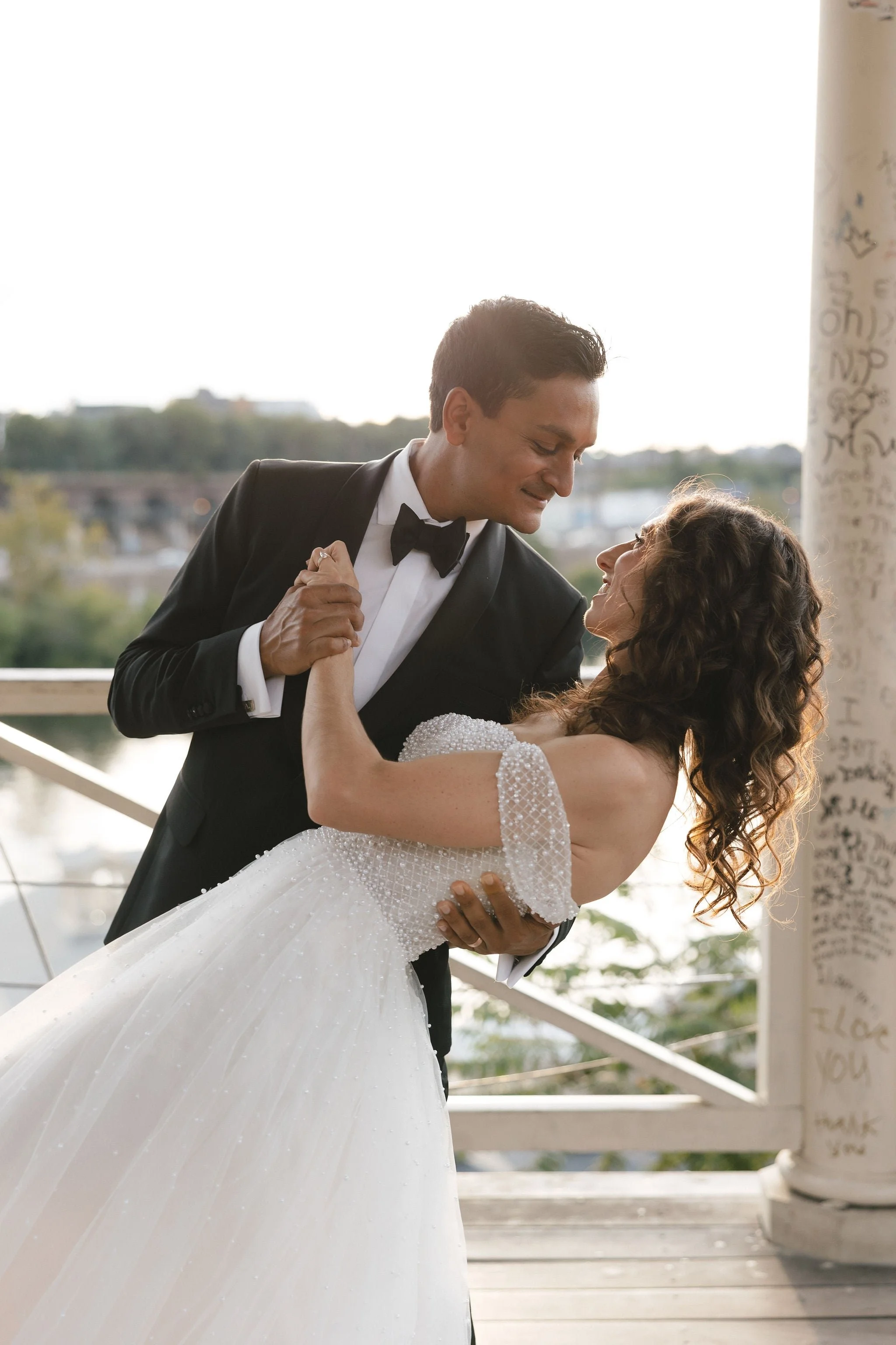 A man in a tuxedo dancing with a woman in a wedding gown on a wooden deck, with an outdoor cityscape in the background and sunlight behind them.