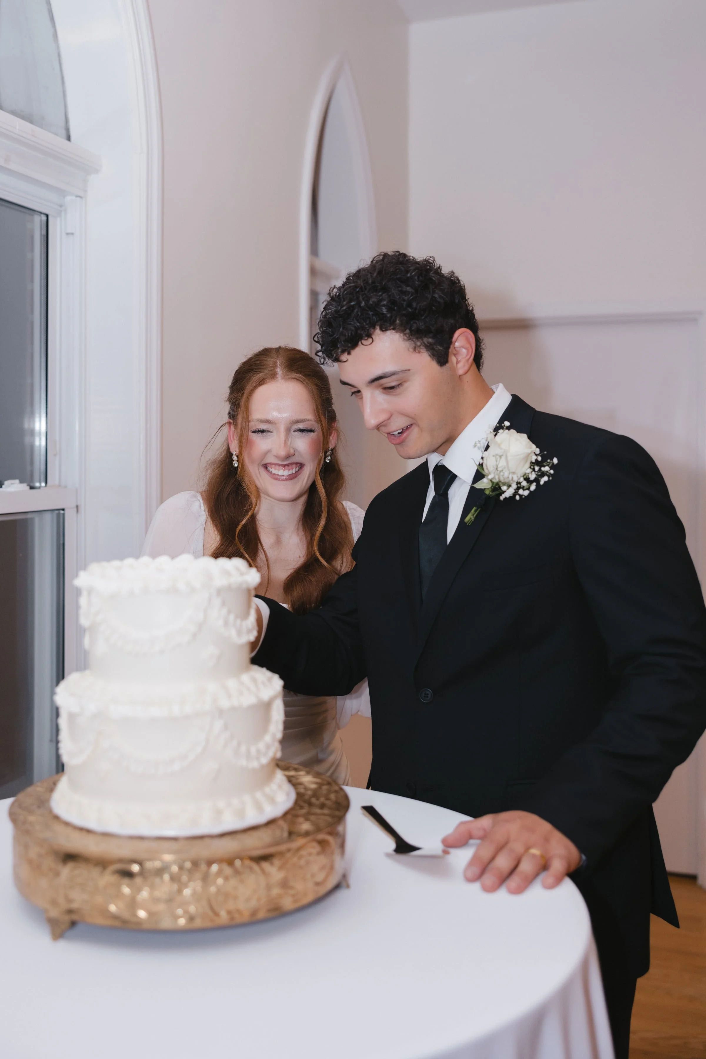 A bride and groom cutting a wedding cake together at their wedding reception. The bride has long red hair and is smiling, wearing pearl earrings and a white dress. The groom has dark curly hair, wearing a black tuxedo with a white flower boutonniere.