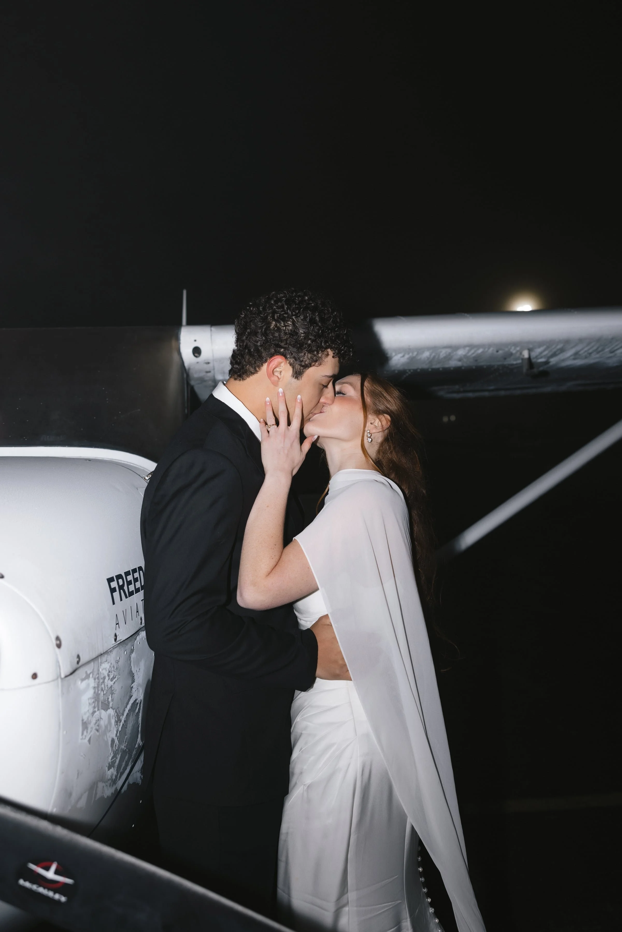 A bride and groom sharing a kiss in front of a small airplane at night.