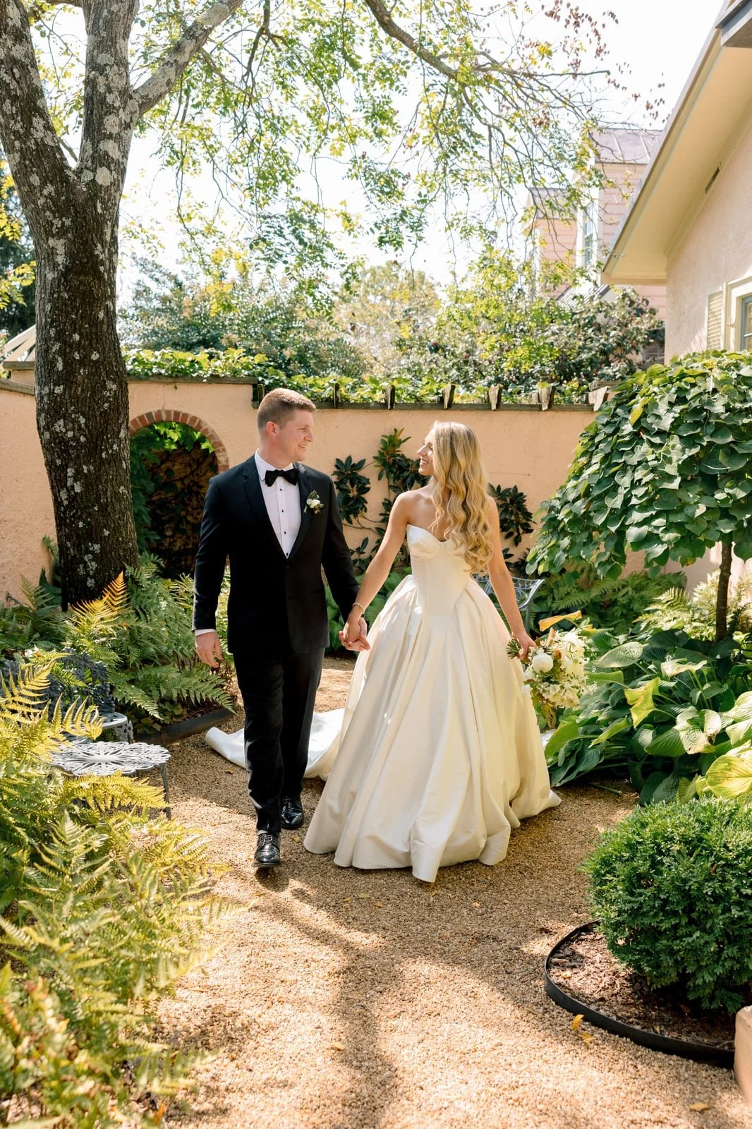 A bride and groom walking hand in hand in a garden, smiling at each other. The bride is wearing a strapless white wedding gown and holding a bouquet, while the groom is dressed in a black tuxedo with a bow tie. The garden has lush green plants, trees