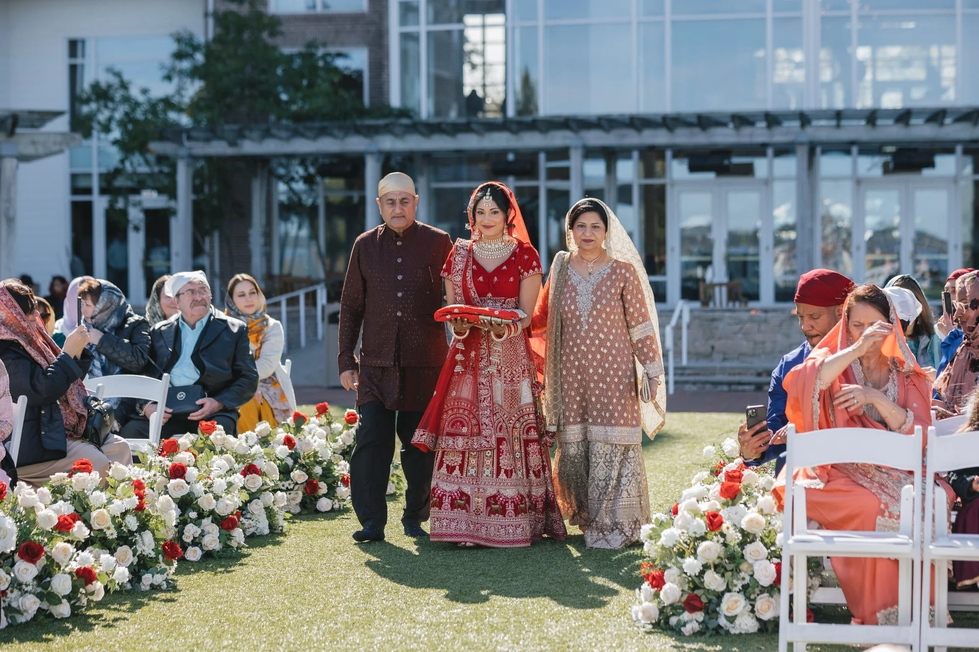 A bride in a red traditional dress with jewelry walking with an older man and woman at an outdoor wedding ceremony. Guests are seated on white chairs, with some taking photos, and decorated with flowers.