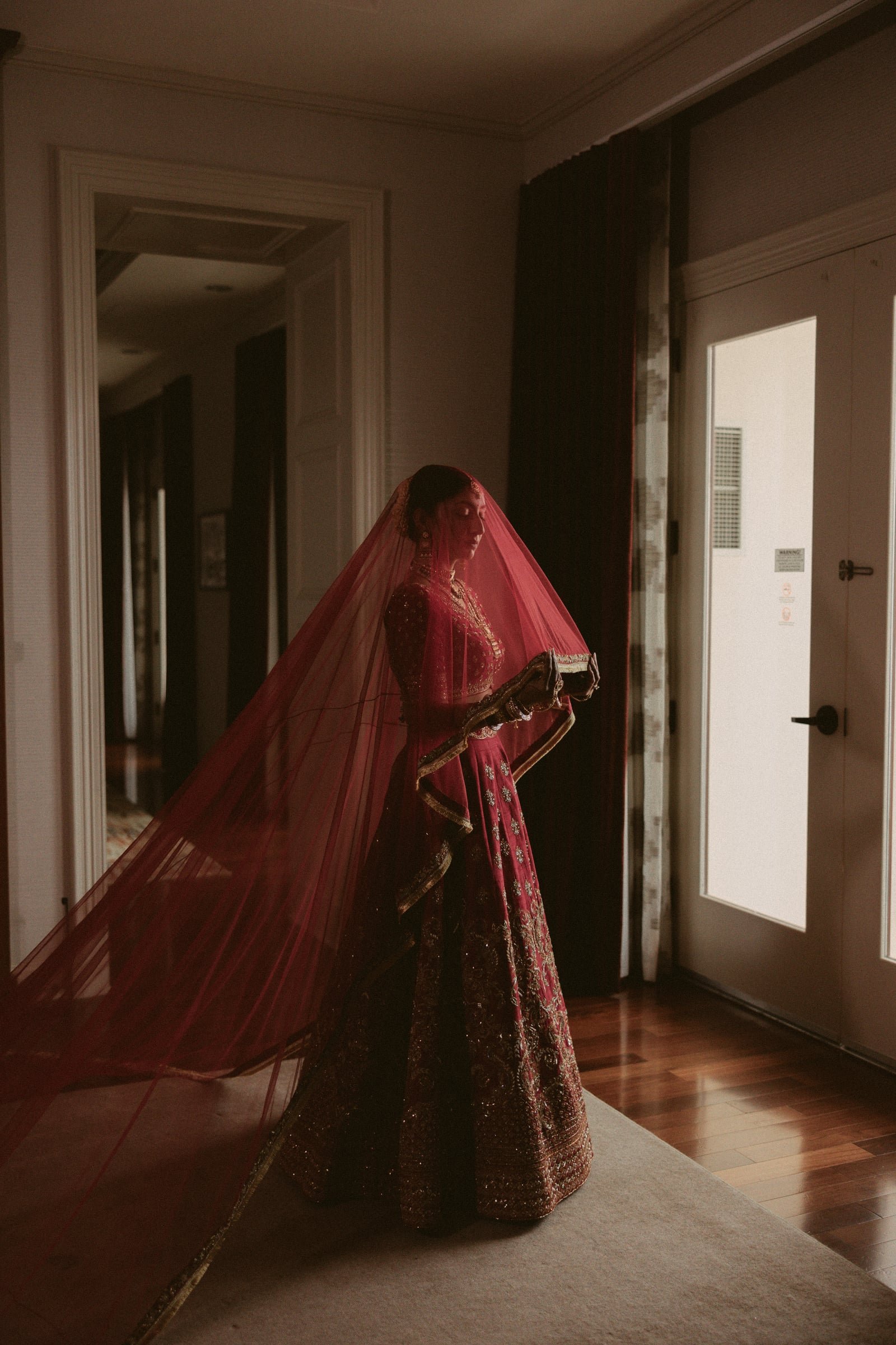 A woman in traditional Indian bridal attire, wearing a red and gold lehenga with intricate embroidery, stands indoors near a door with glass panels, partially covered by a long red veil. The room has wooden flooring, a beige carpet, and dark curtains