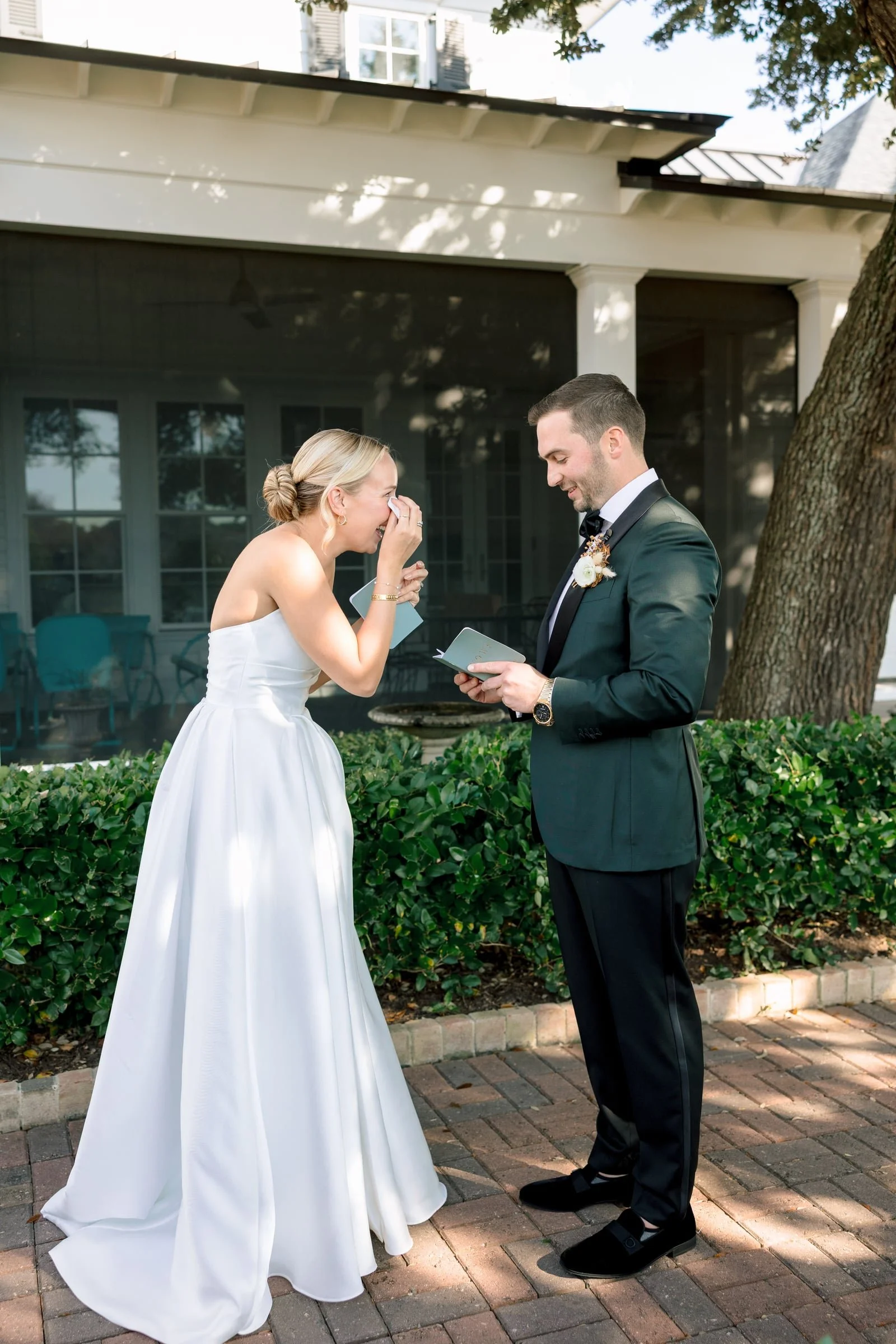 A bride and groom stand outdoors in front of a house, dressed in wedding attire. The bride is wearing a white strapless wedding gown and is laughing, covering her mouth with her hand. The groom is wearing a dark suit and a bow tie, holding a small folded paper or card, smiling, and looking at the bride. There are trees, green bushes, and a brick pathway in the background.