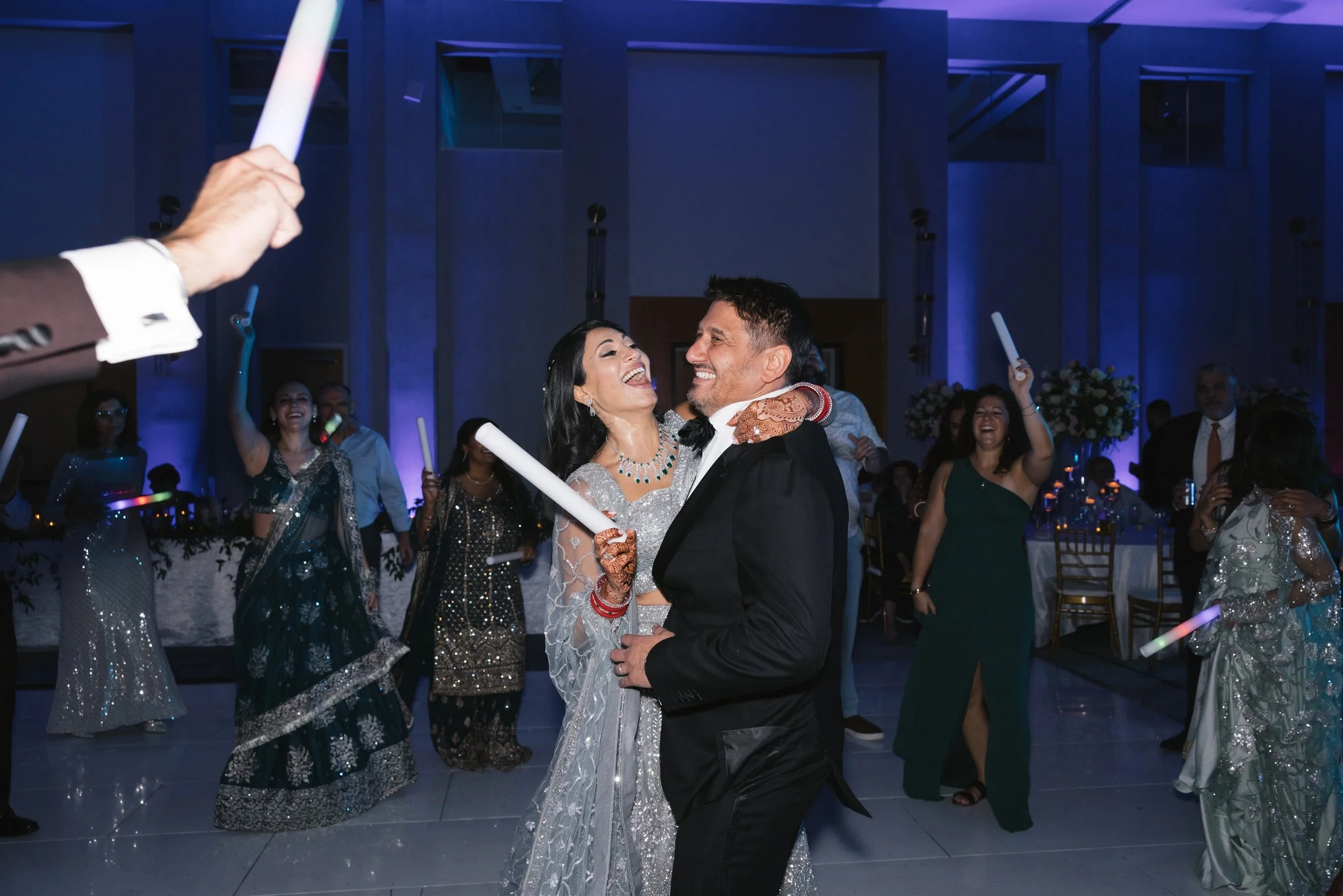 A bride and groom dancing together at a wedding reception surrounded by guests, with the bride wearing a silver dress and jewelry and the groom in a black tuxedo, all smiling and enjoying the celebration.