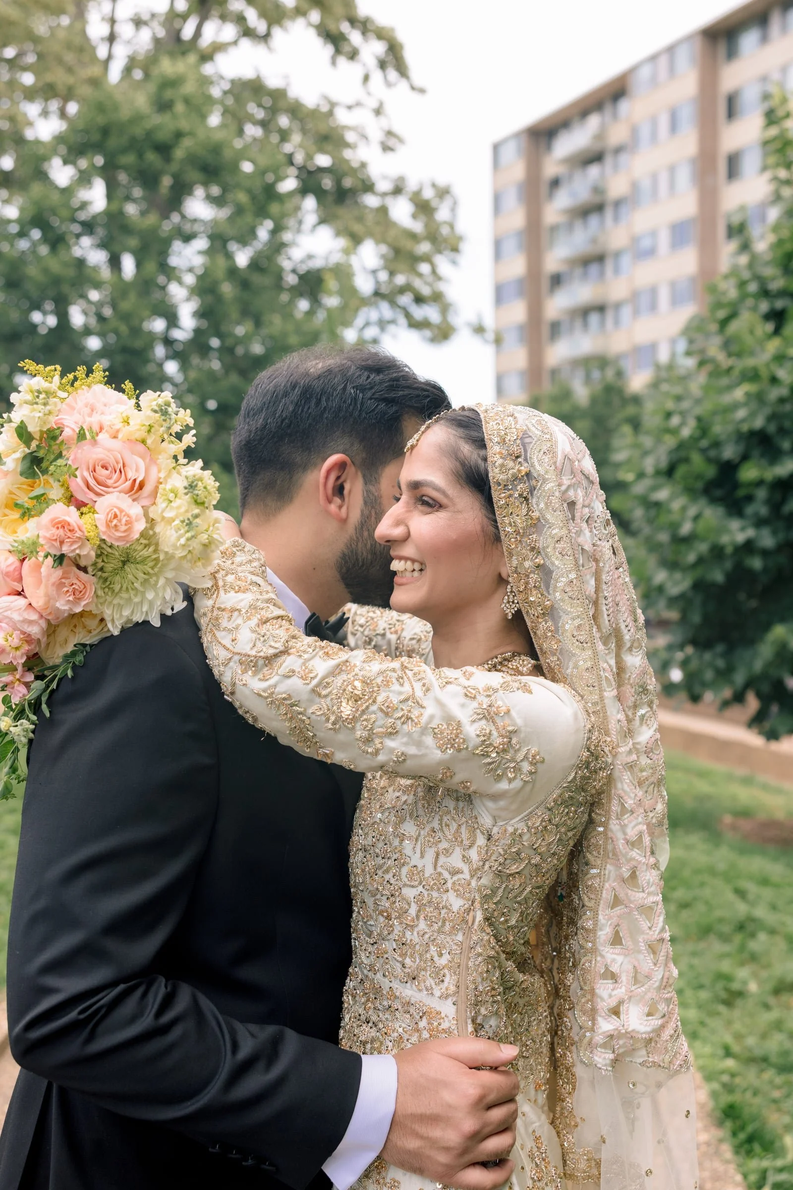 A bride and groom embrace outdoors during their wedding. The bride wears an ornate gold and cream dress with a head covering, and the groom is dressed in a black suit and bow tie. The bride holds a large bouquet of pink and white flowers, and they ar