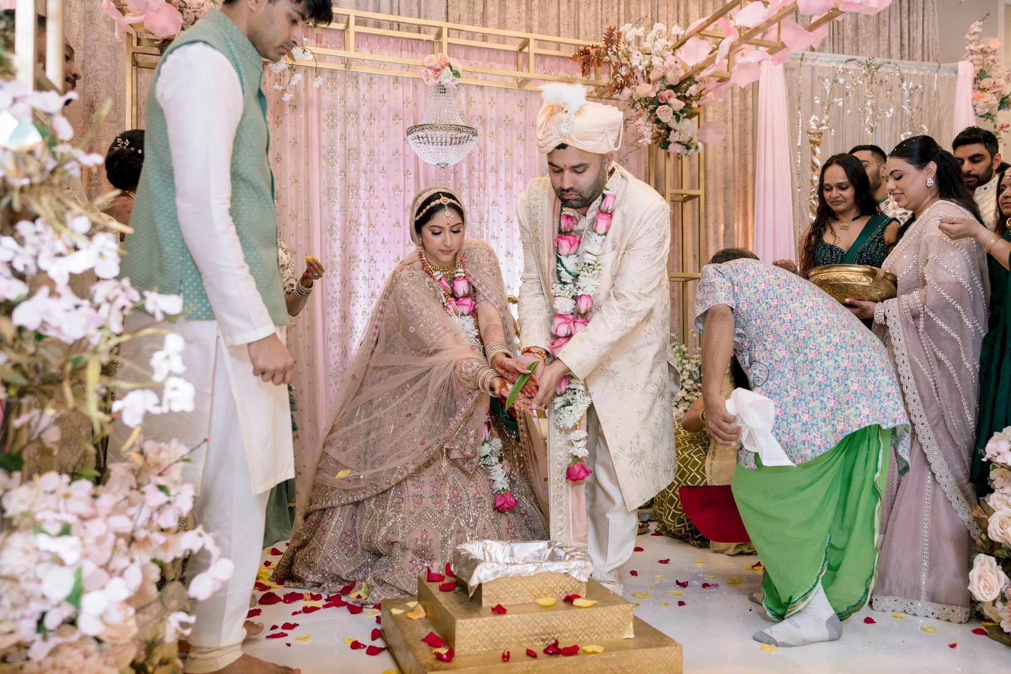 A traditional Indian wedding ceremony, with the bride and groom performing a ritual, surrounded by family and friends in a decorated venue with floral arrangements and drapes.
