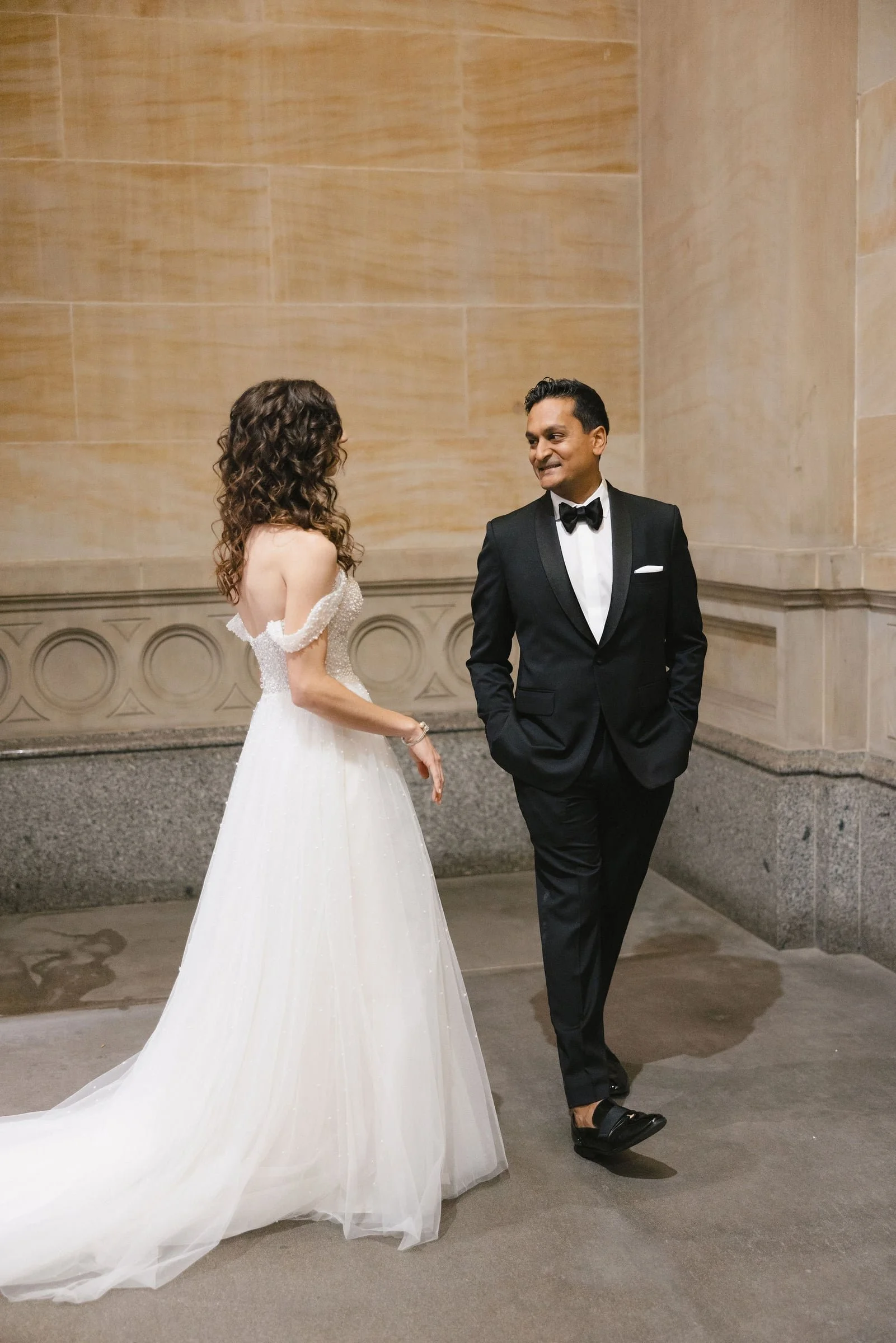 A bride and groom dressed in wedding attire standing against a beige stone wall, smiling and looking at each other.