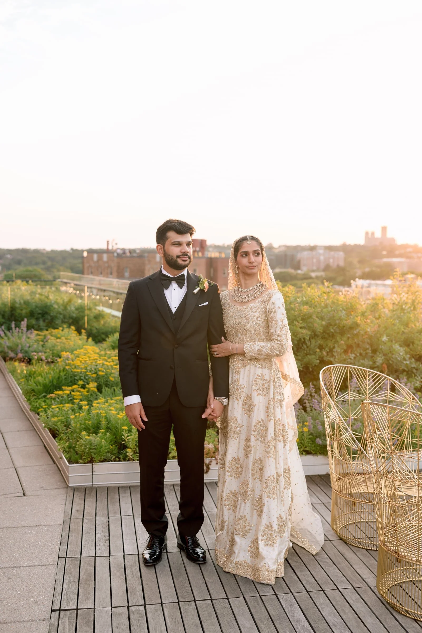 A couple dressed in formal wedding attire, holding hands on a rooftop garden during sunset. The man is wearing a black tuxedo with a bow tie and boutonniere, and the woman is in an elaborate cream-colored dress with gold embroidery and jewelry. There