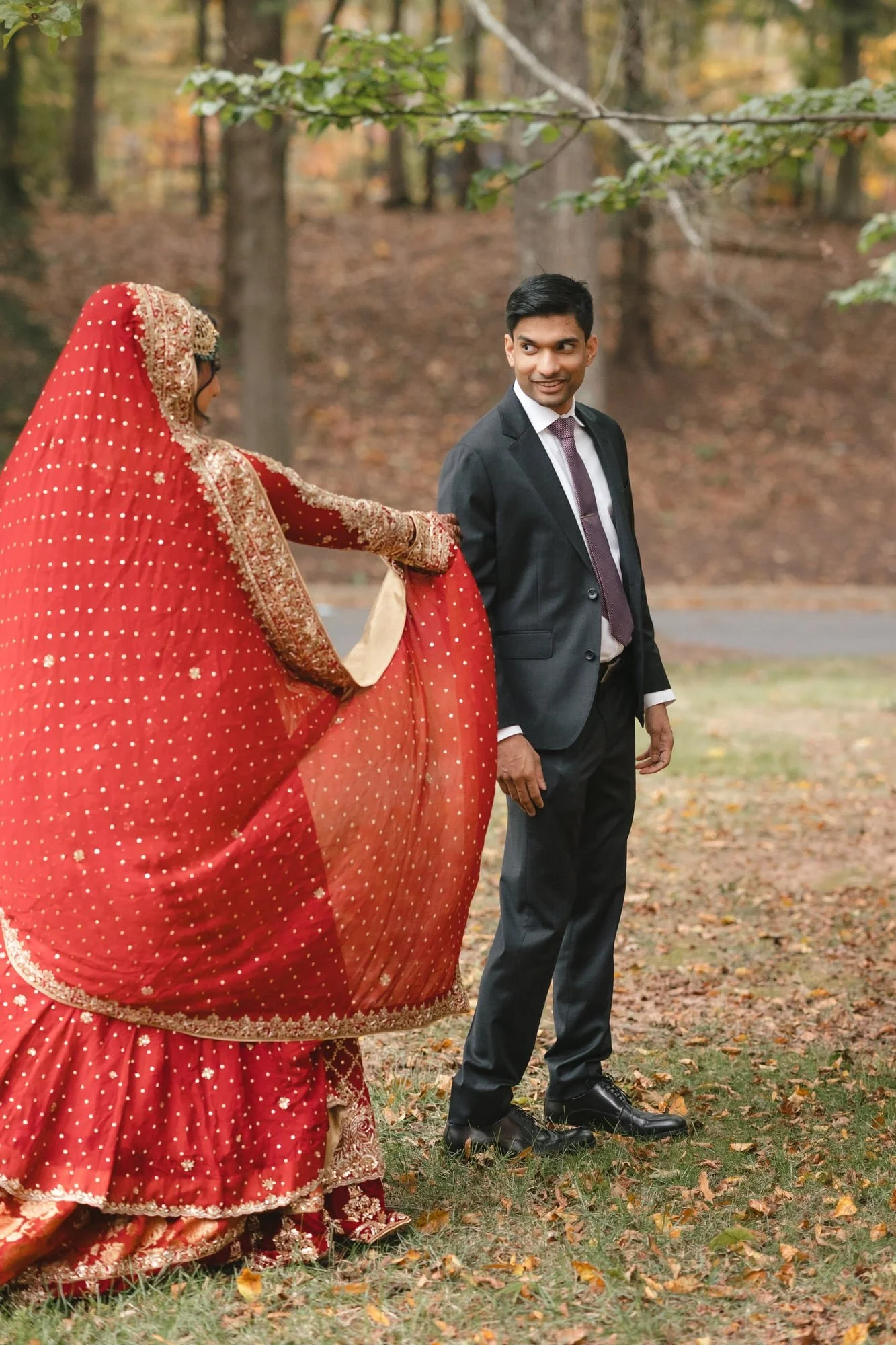 A man in a black suit and tie standing outdoors on fallen autumn leaves, smiling while holding hands with a woman dressed in a red traditional South Asian outfit with gold embroidery.