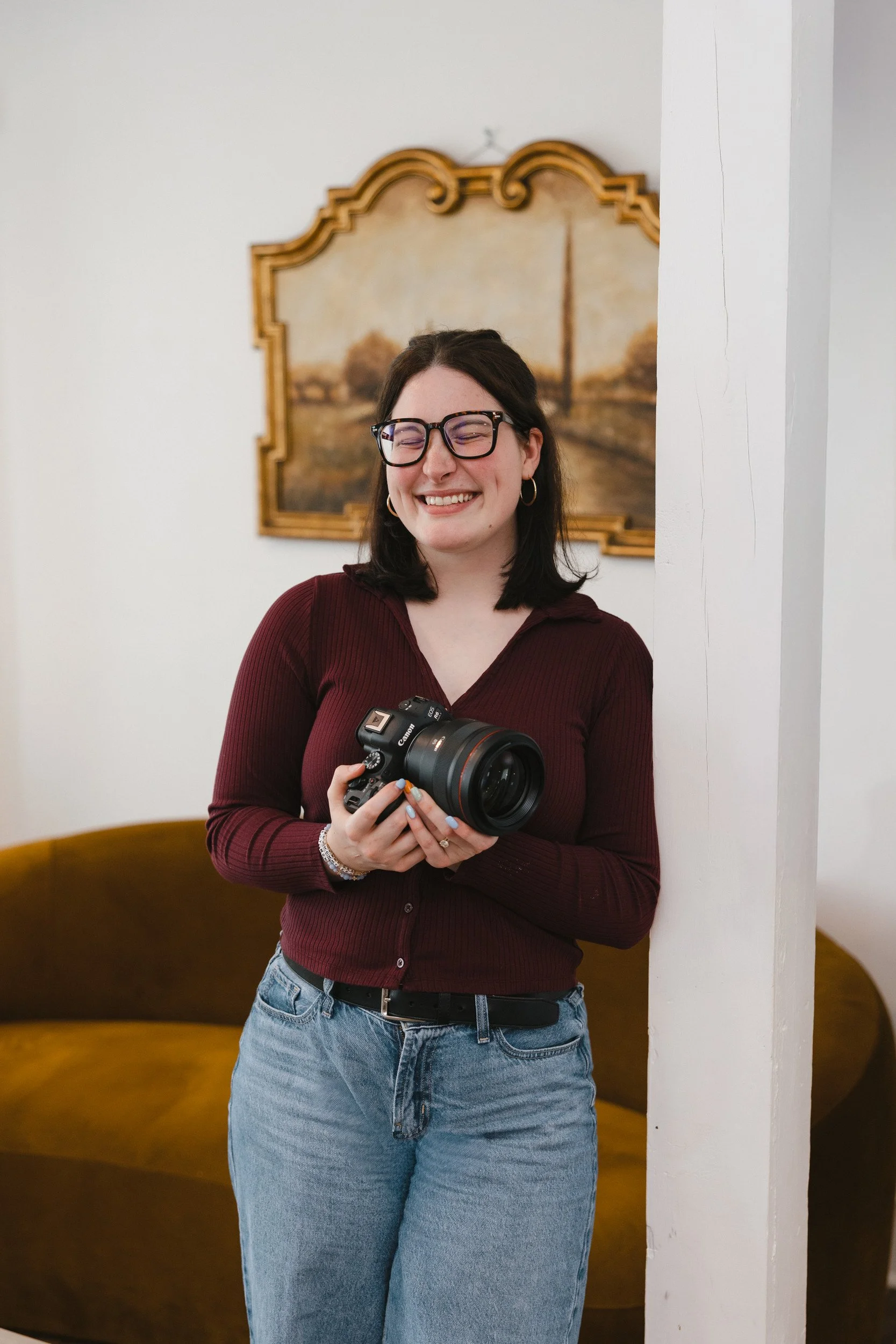 A woman with dark hair, glasses, wearing a burgundy long-sleeve shirt and blue jeans, holding a camera and smiling, standing beside a white wall with a painting behind her
