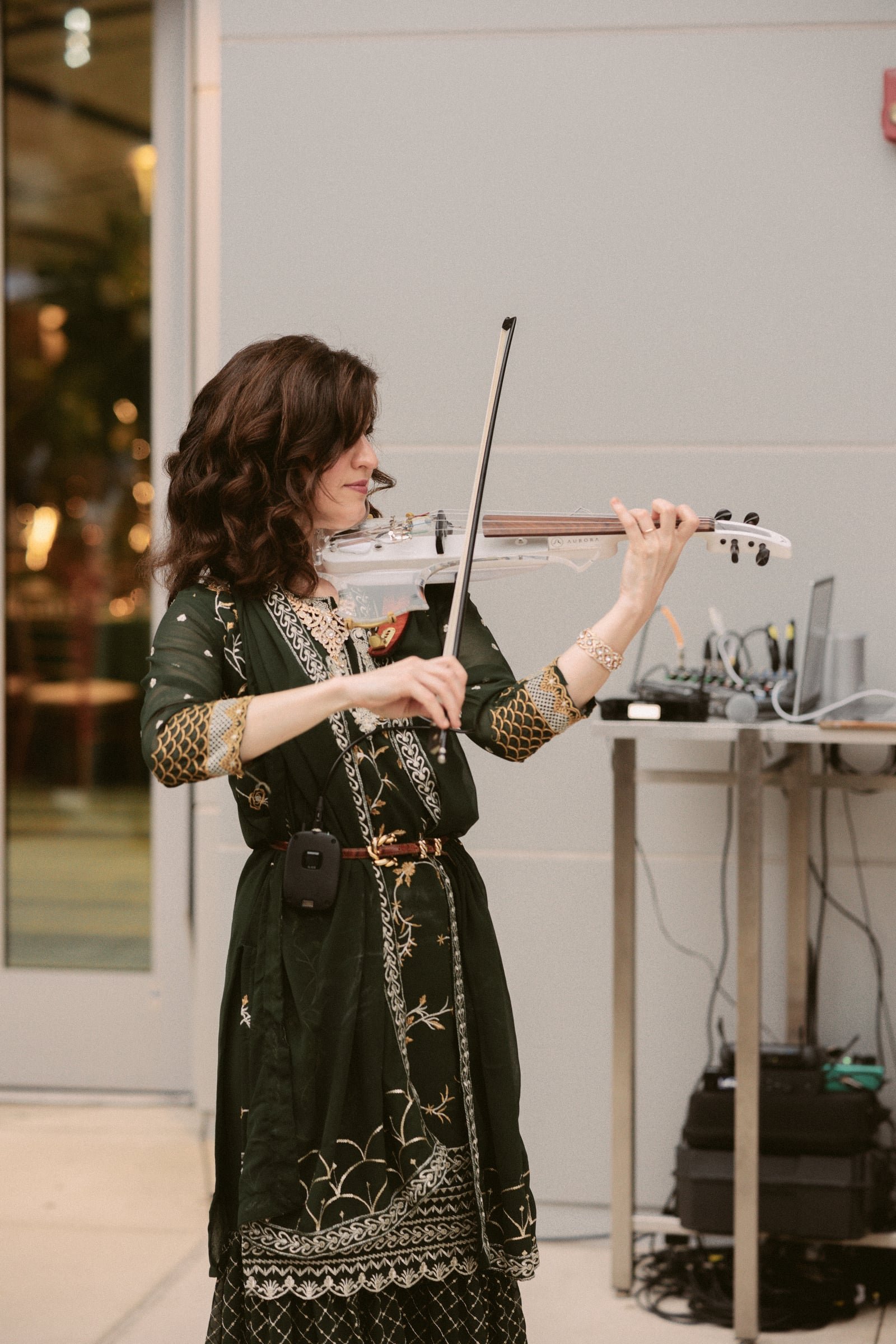 A woman with curly brown hair wearing a black and gold embroidered dress playing a white electric violin in an indoor setting.