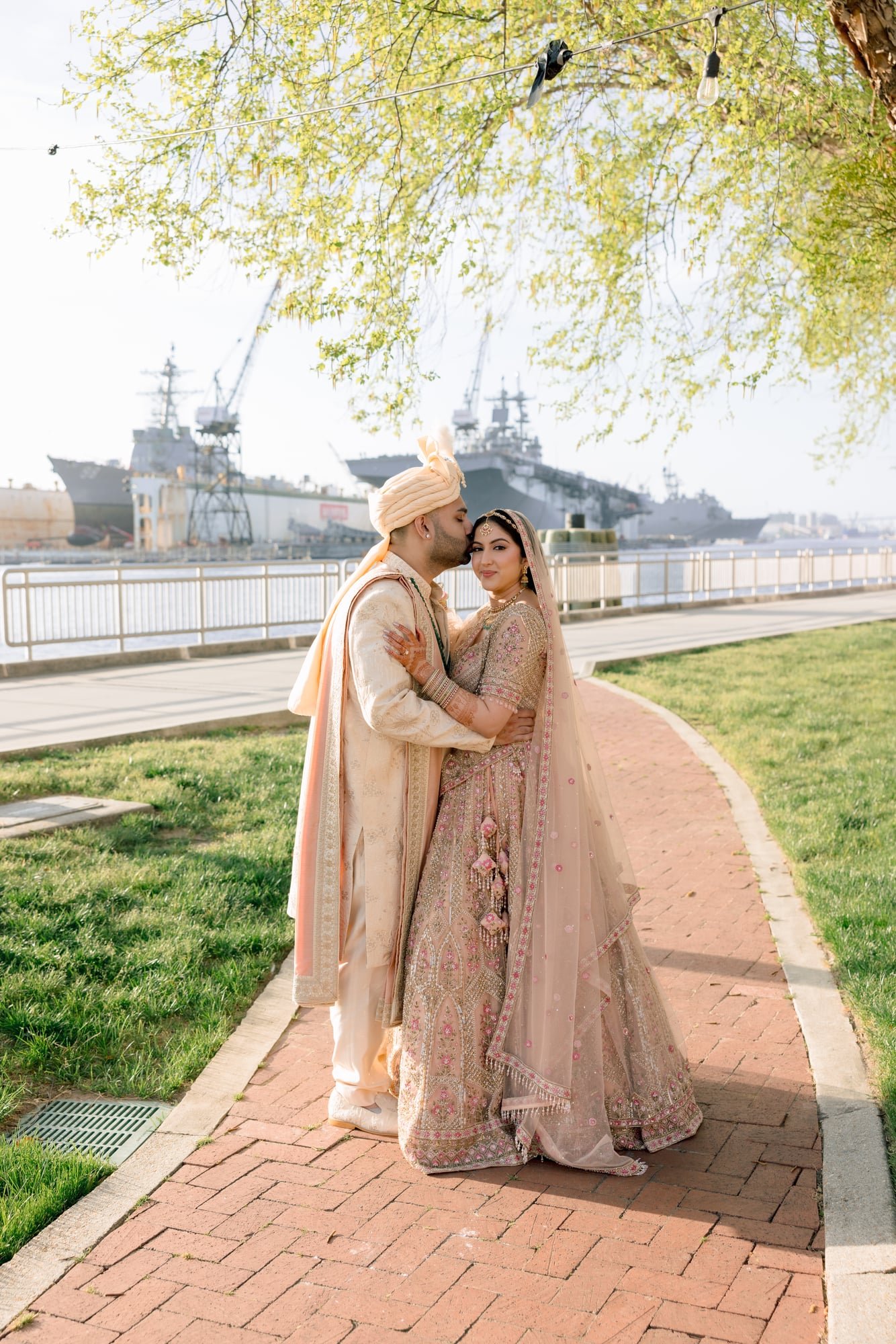 A newlywed South Asian couple in traditional wedding attire standing close, outdoors near a waterfront, with large ships, cranes, and trees in the background.