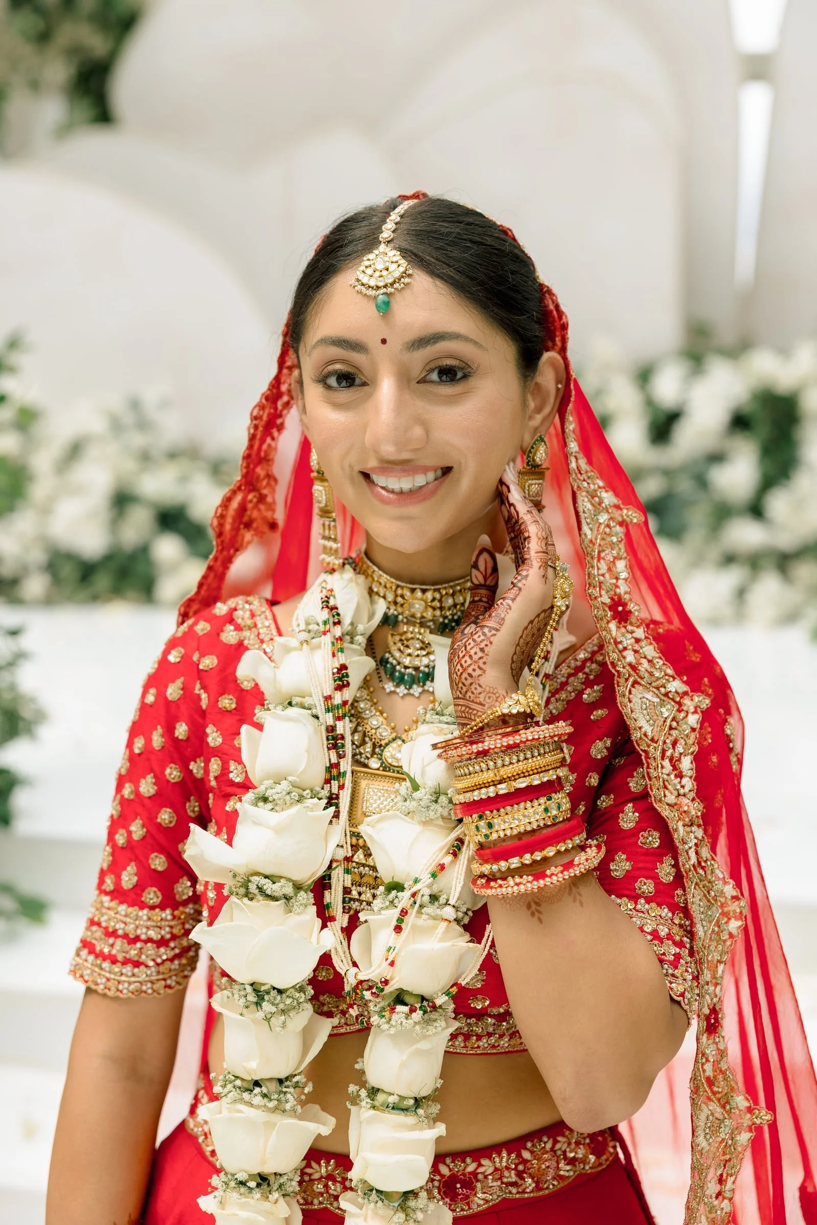 A woman dressed in traditional Indian bridal attire, wearing red and gold embroidery, ornate jewelry including a maang tikka, earrings, necklace, and bangles, standing with a floral garland of white flowers around her neck, smiling at the camera.
