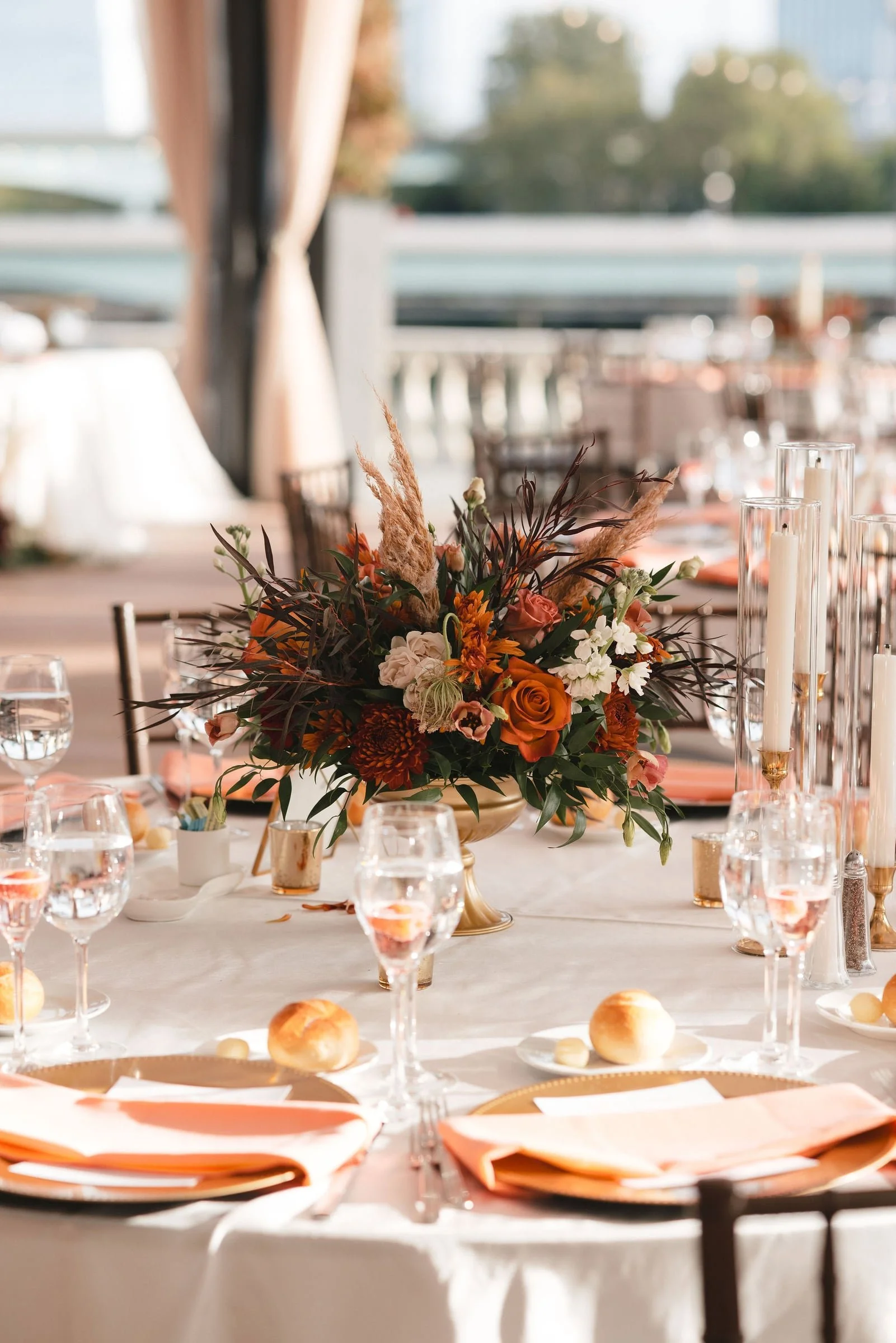 A round table set for a formal event with a centerpiece of orange, white, and brown flowers, tall glass candle holders with white candles, glasses of water, and small bread rolls on white plates with peach napkins. The background shows a bright, sunl