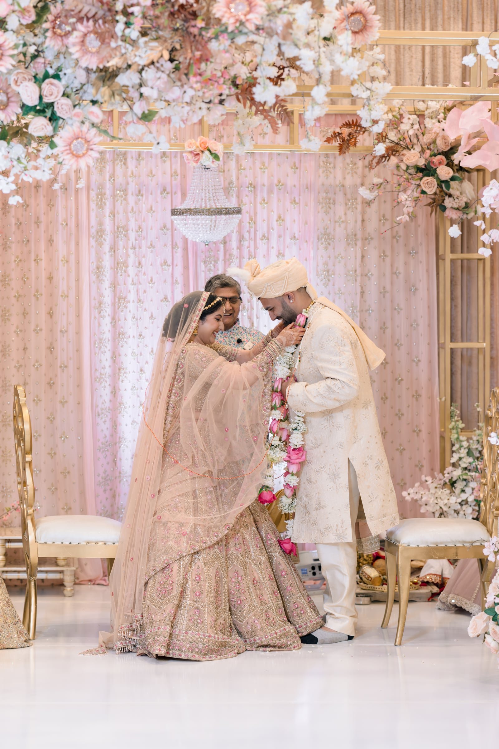 Indian bride and groom smiling during their wedding ceremony, surrounded by pink and white floral decorations and a chandelier.
