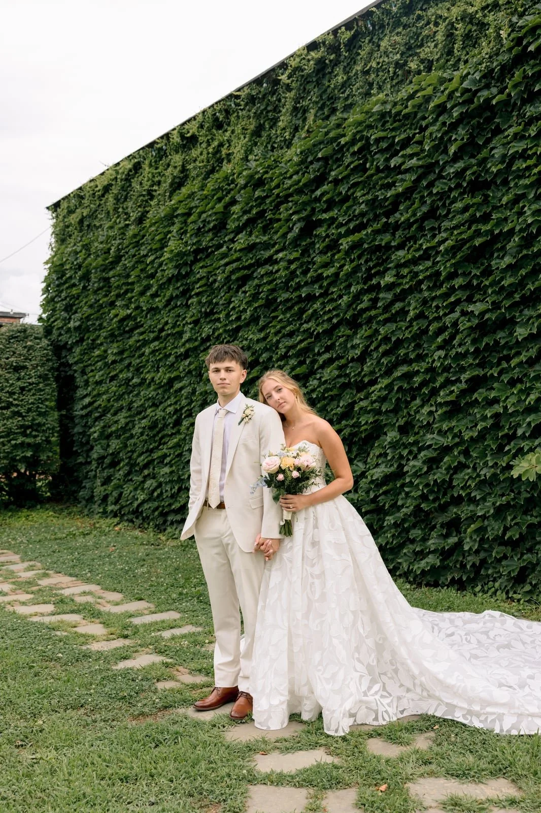 A young couple in wedding attire holding hands outdoors next to a large green leafy wall, with the woman holding a bouquet of flowers.