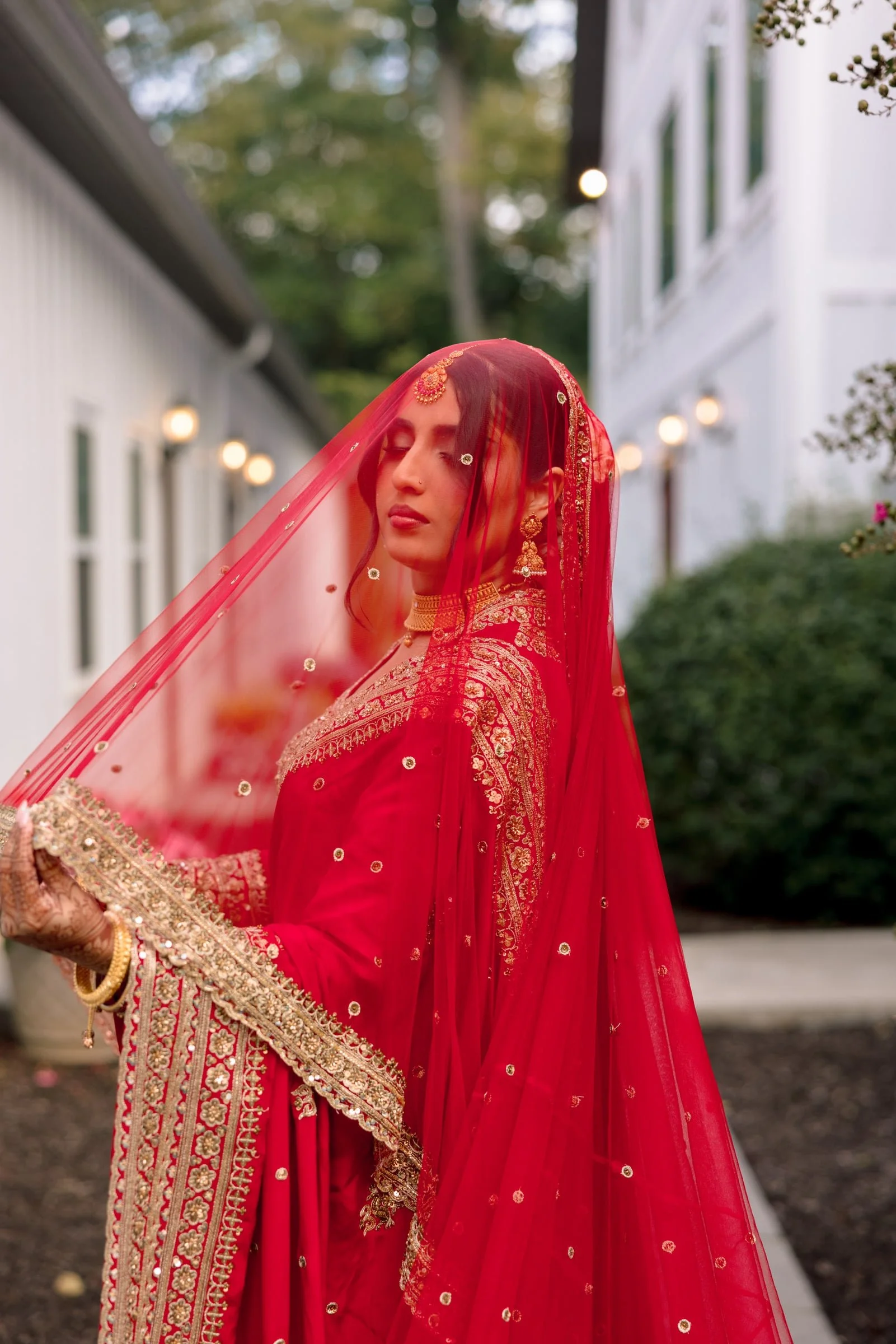 A woman wearing a red traditional dress with intricate gold embroidery, and a red veil with small mirrors, standing outdoors with closed eyes.