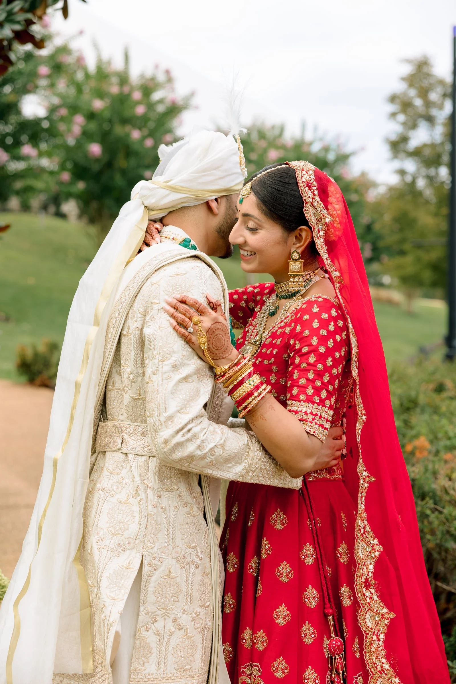 A bride and groom dressed in traditional Indian wedding attire sharing a close and joyful moment outdoors, with trees and a cloudy sky in the background.