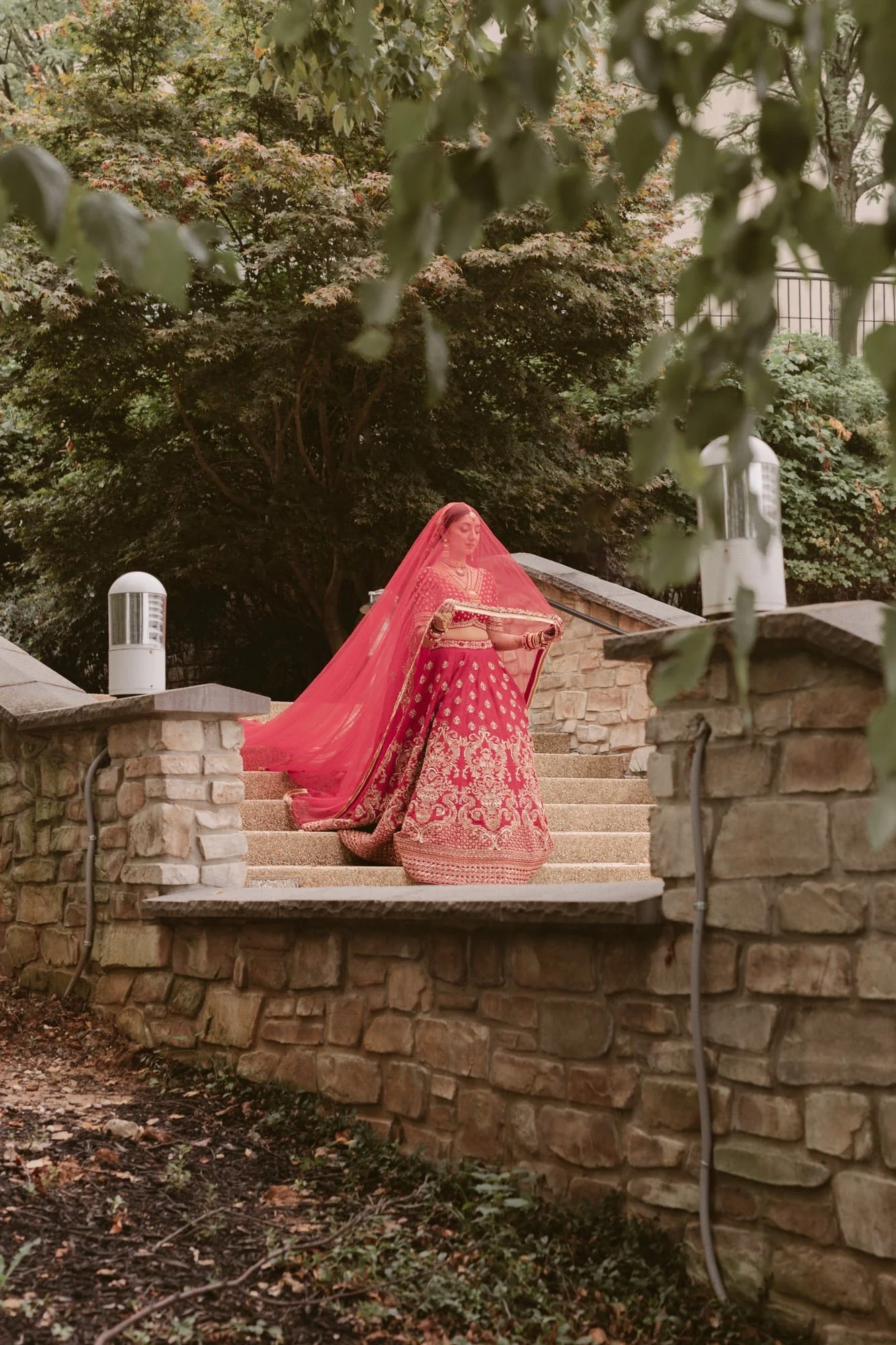 A woman dressed in a traditional pink and gold Indian saree with jewelry, standing on outdoor stone steps, with a pink dupatta draped over her head, holding a sword, surrounded by lush trees and greenery.