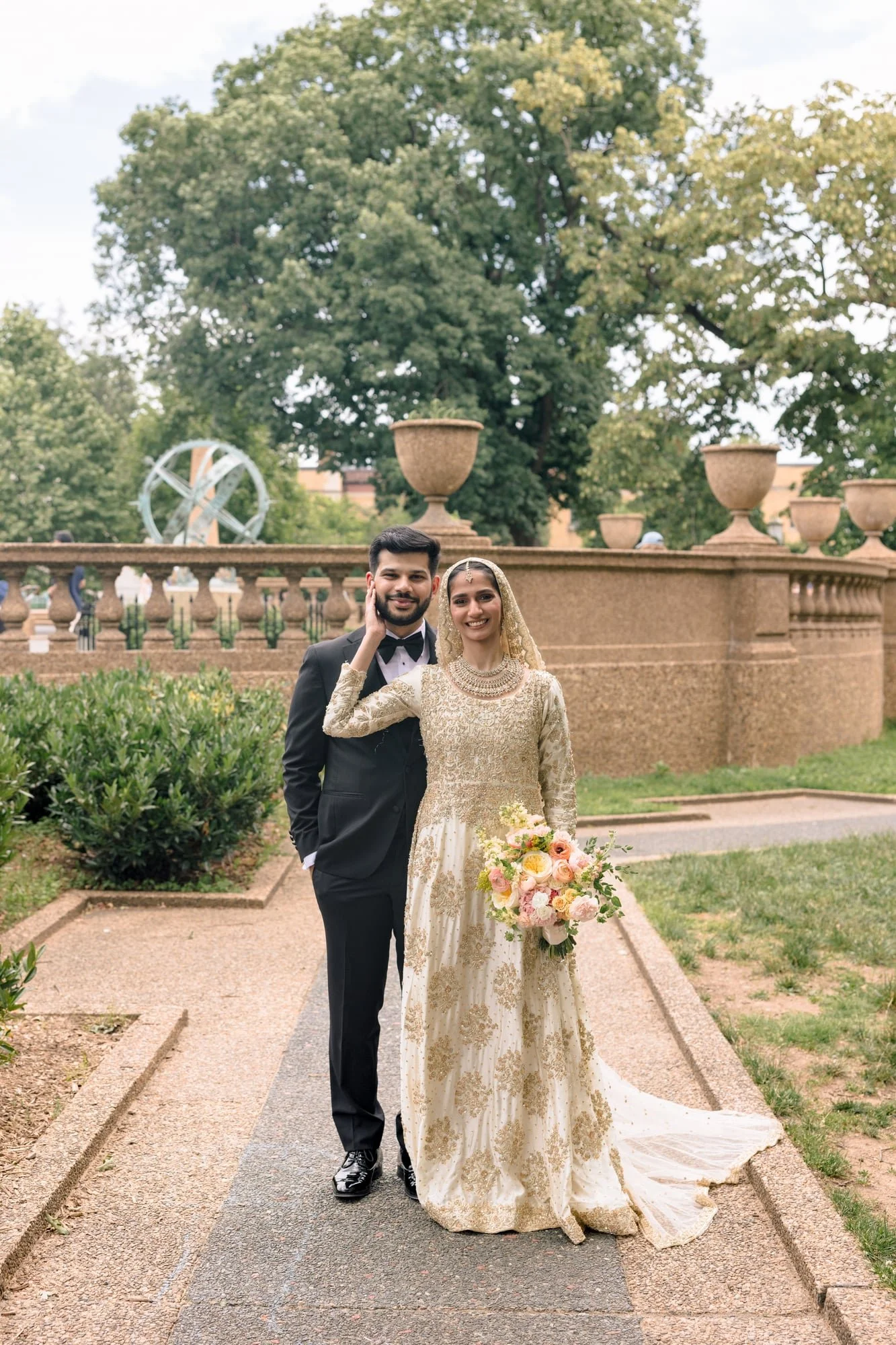 A newlywed couple stands on a garden pathway, smiling at the camera. The groom is dressed in a black tuxedo with a bow tie, and the bride is wearing a gold, embroidered wedding gown with a lace veil and holding a large bouquet of pastel roses and greenery. Behind them is a brick fountain, trees, and a decorative globe sculpture.