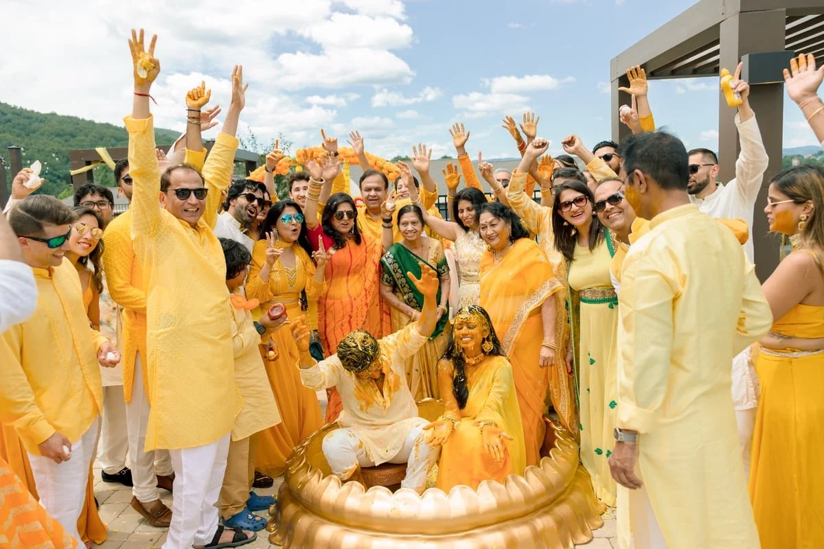 Celebration of an Indian wedding with guests dressed in yellow and orange clothes, some with colorful accessories, gathered around the bride and groom who are seated and covered in yellow powder, indicating a traditional pre-wedding ritual outdoors in sunny weather.