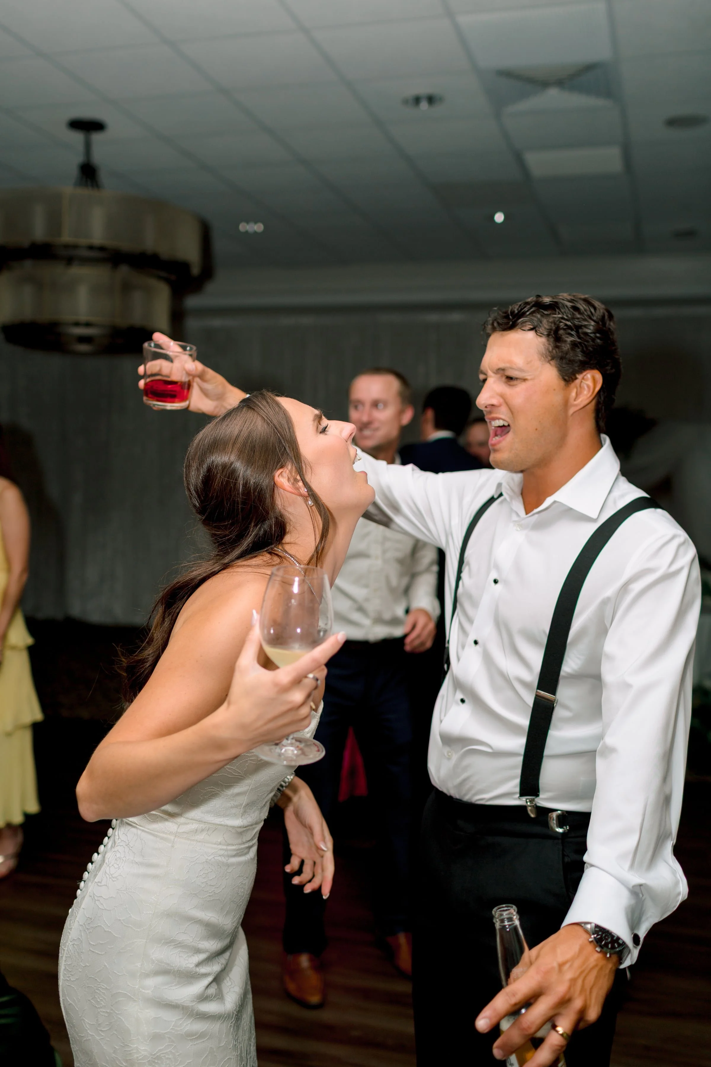 A man and woman dancing together at a party, holding drinks, with other guests in the background.