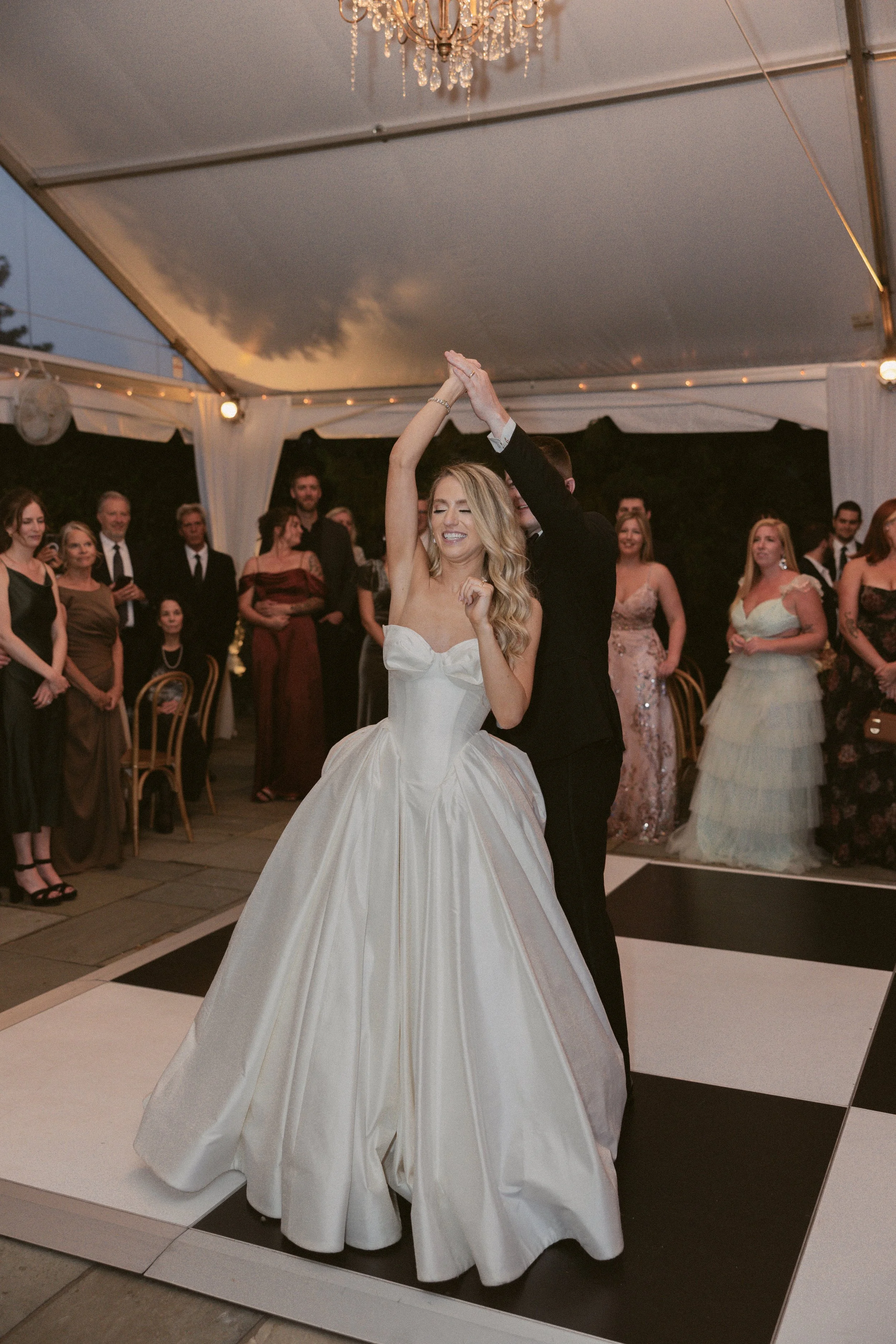 A bride and groom dance at their wedding reception under a white tent with a chandelier. Guests in formal attire watch the couple, with some smiling and some taking photos.