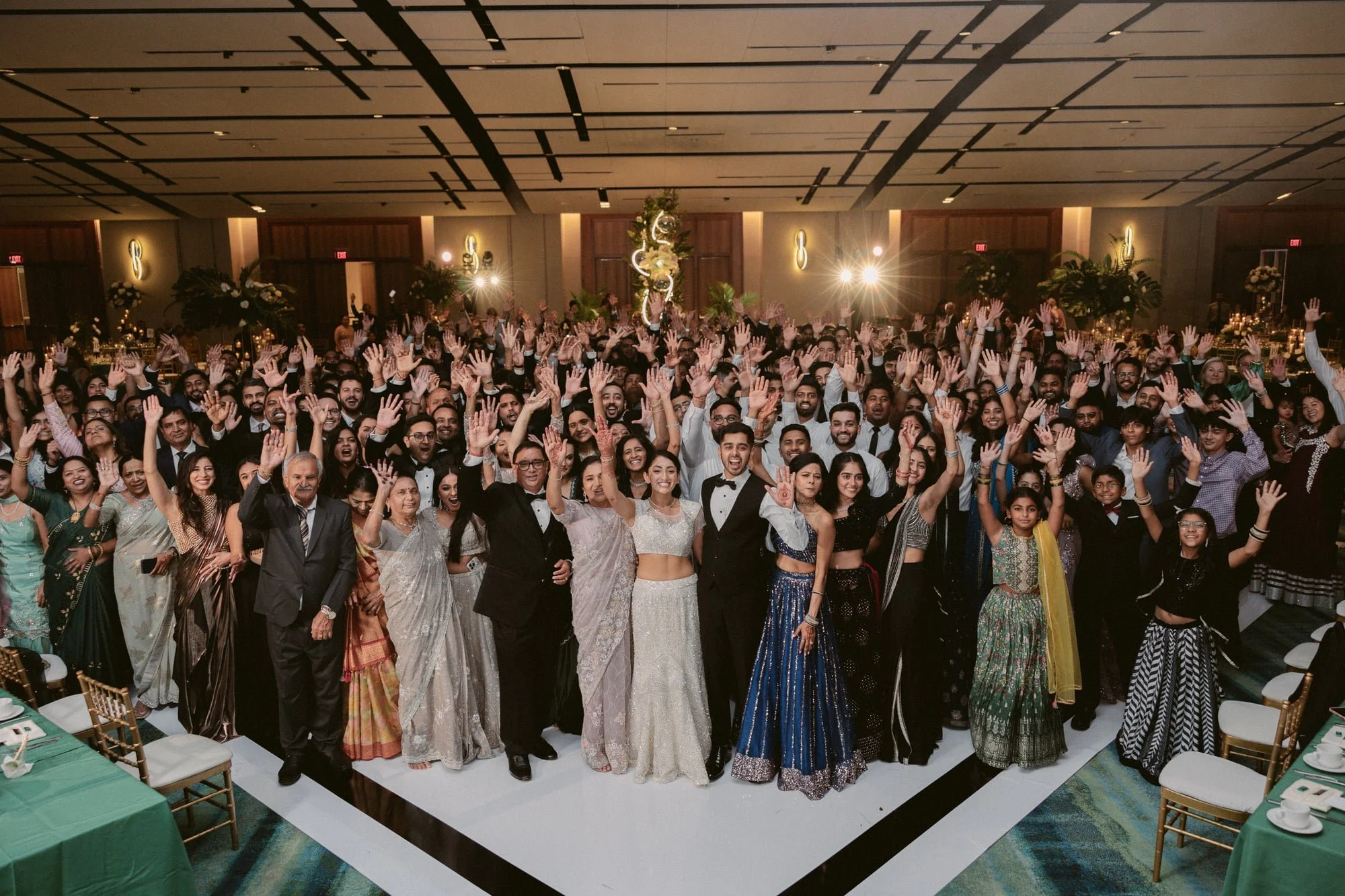 A large group of people dressed in traditional Indian attire and western tuxedos at a banquet hall, raising their hands in celebration or greeting.