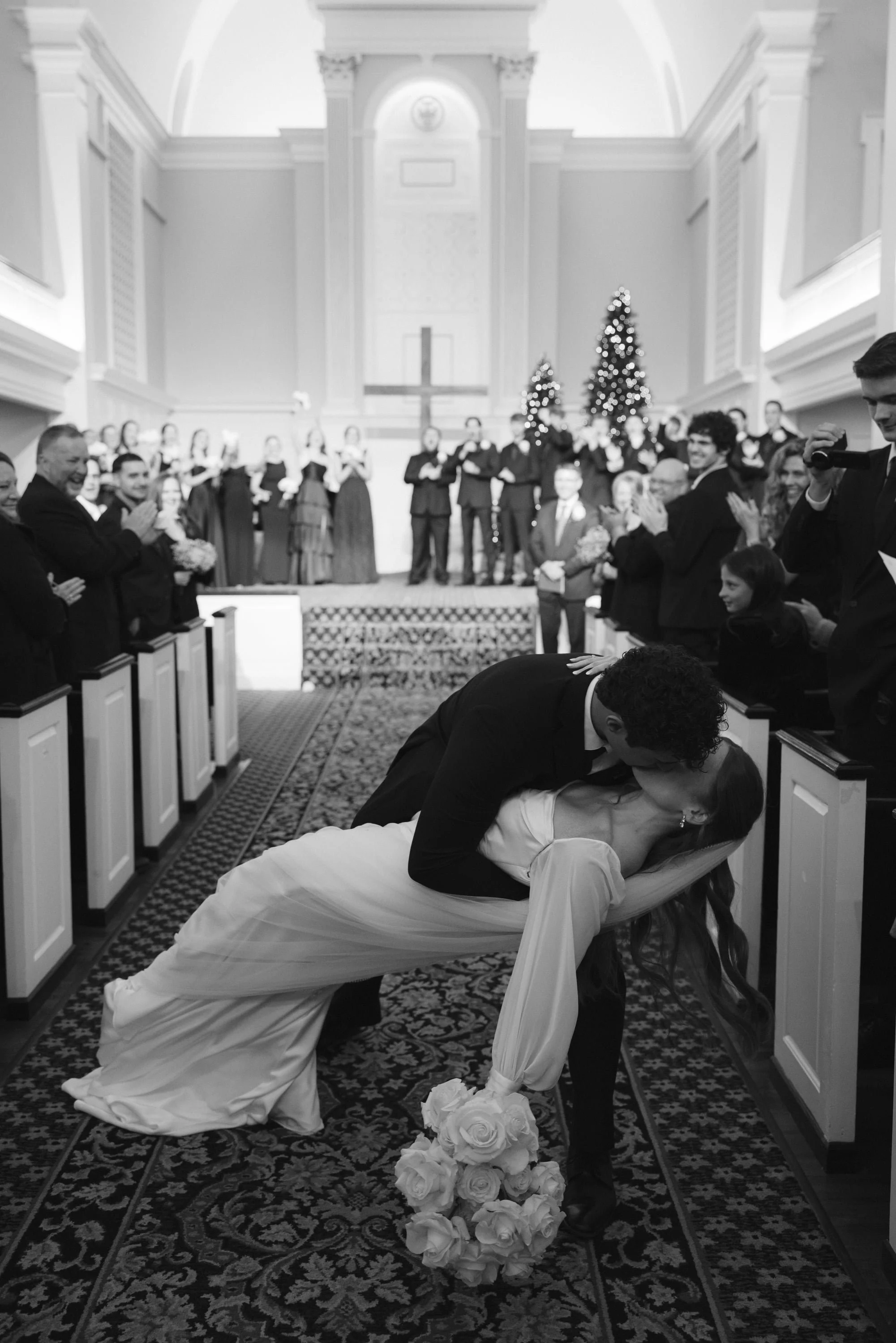 Couple kissing on their wedding day in a church, with a wedding arch, flowers, and a choir in the background.