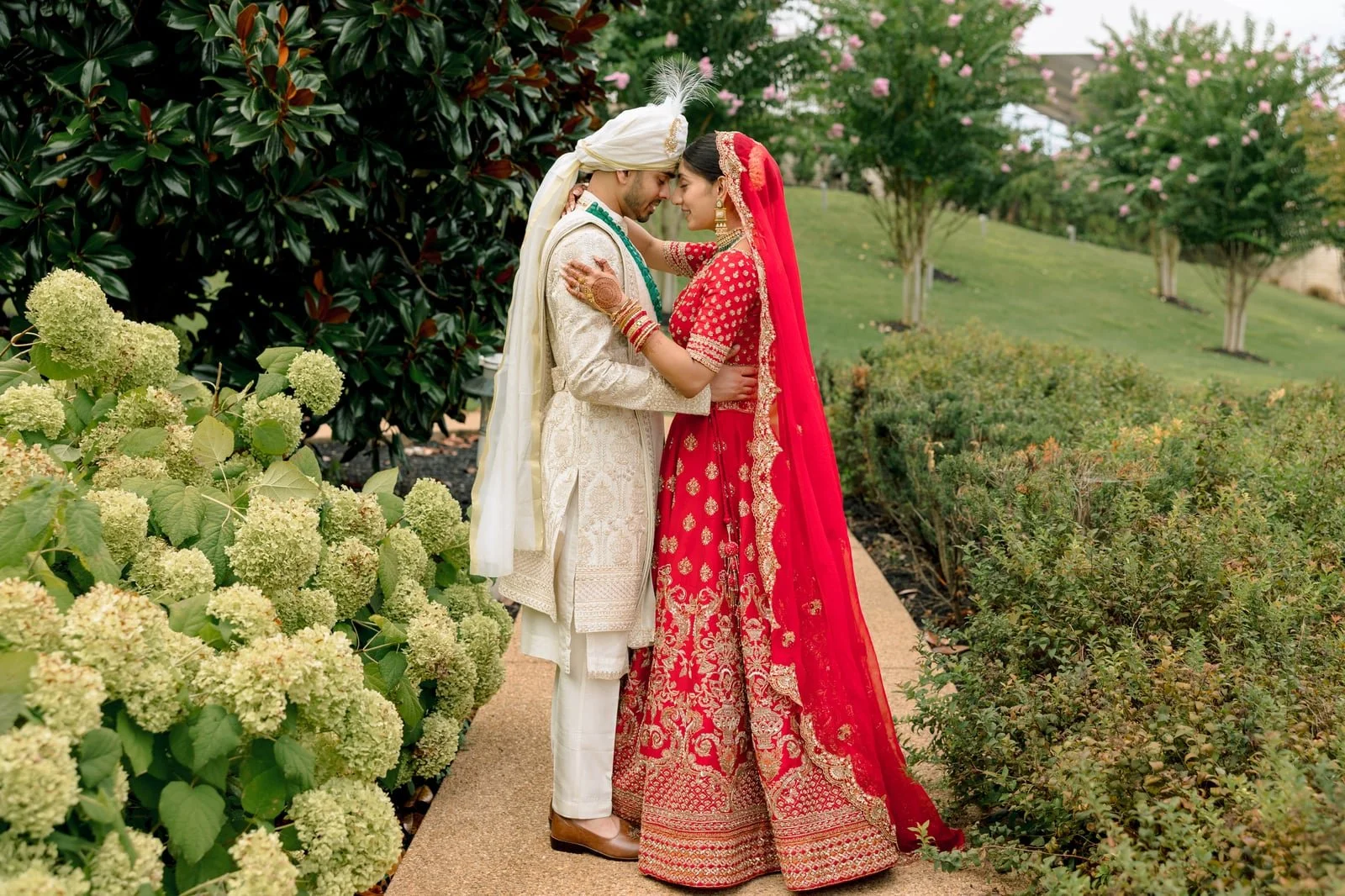 A couple in traditional Indian wedding attire embracing outdoors on a garden pathway, surrounded by flowering plants and trees.