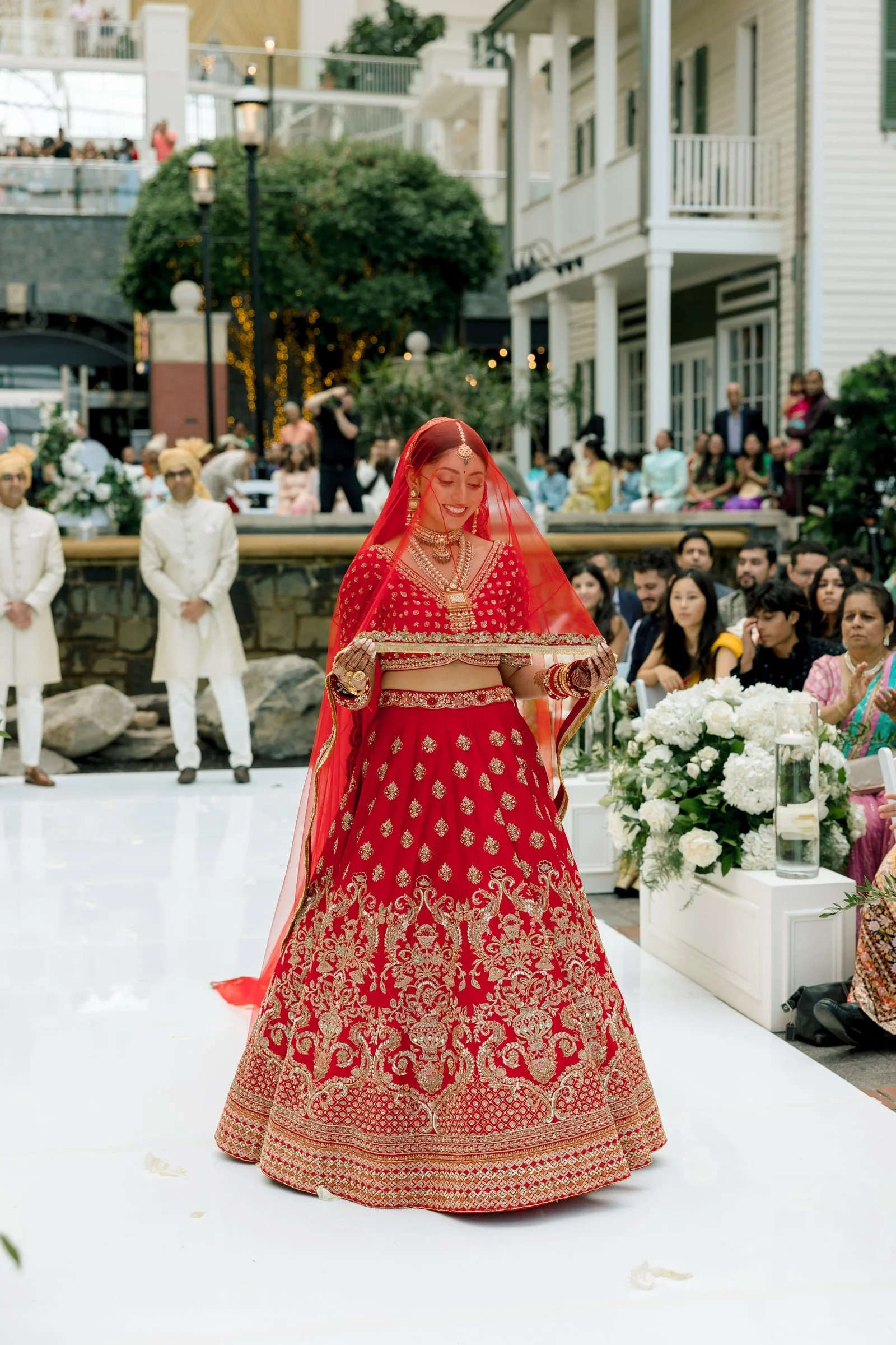 A woman dressed in traditional Indian wedding attire, wearing a red embellished saree with gold embroidery, a matching red veil, and various jewelry, standing and smiling at a wedding ceremony, with guests seated behind her.