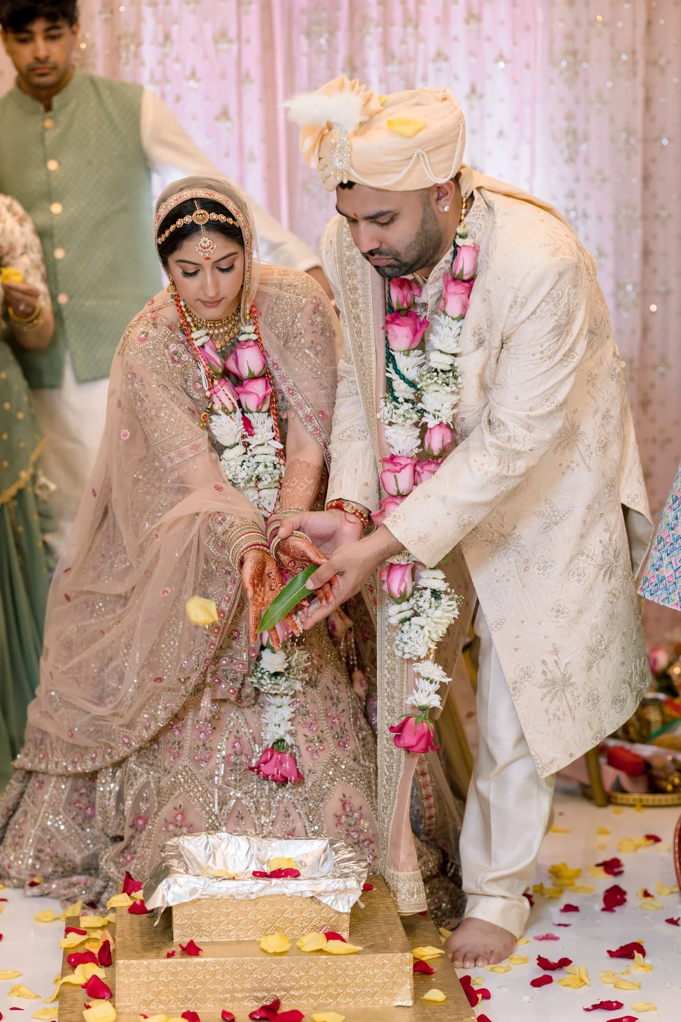 An Indian bride and groom performing a wedding ritual, bowing over a decorated platform with flower petals scattered around, in a traditional wedding setting.