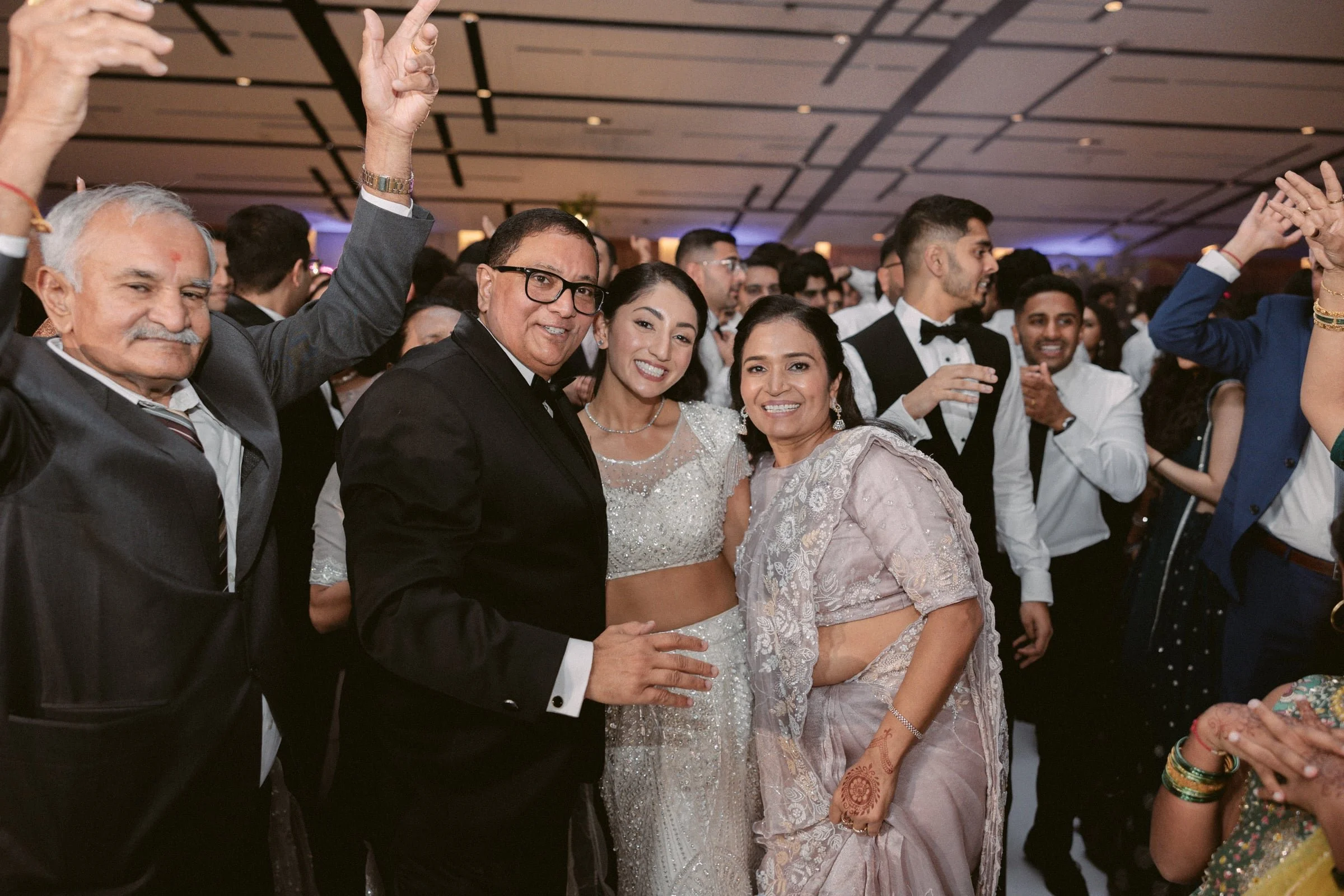 A group of people dressed in formal attire celebrating at a festive event, with some raising their hands and smiling for the camera.