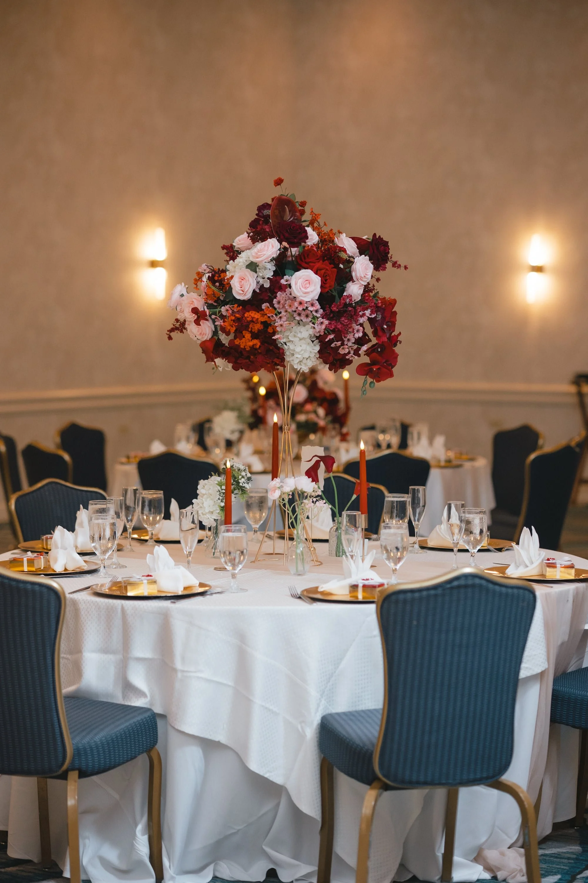 A round banquet table decorated with white tablecloth, floral centerpiece with red, pink, and white flowers, surrounded by blue chairs, in a decorated banquet hall with beige walls and wall sconces, set for a formal event.