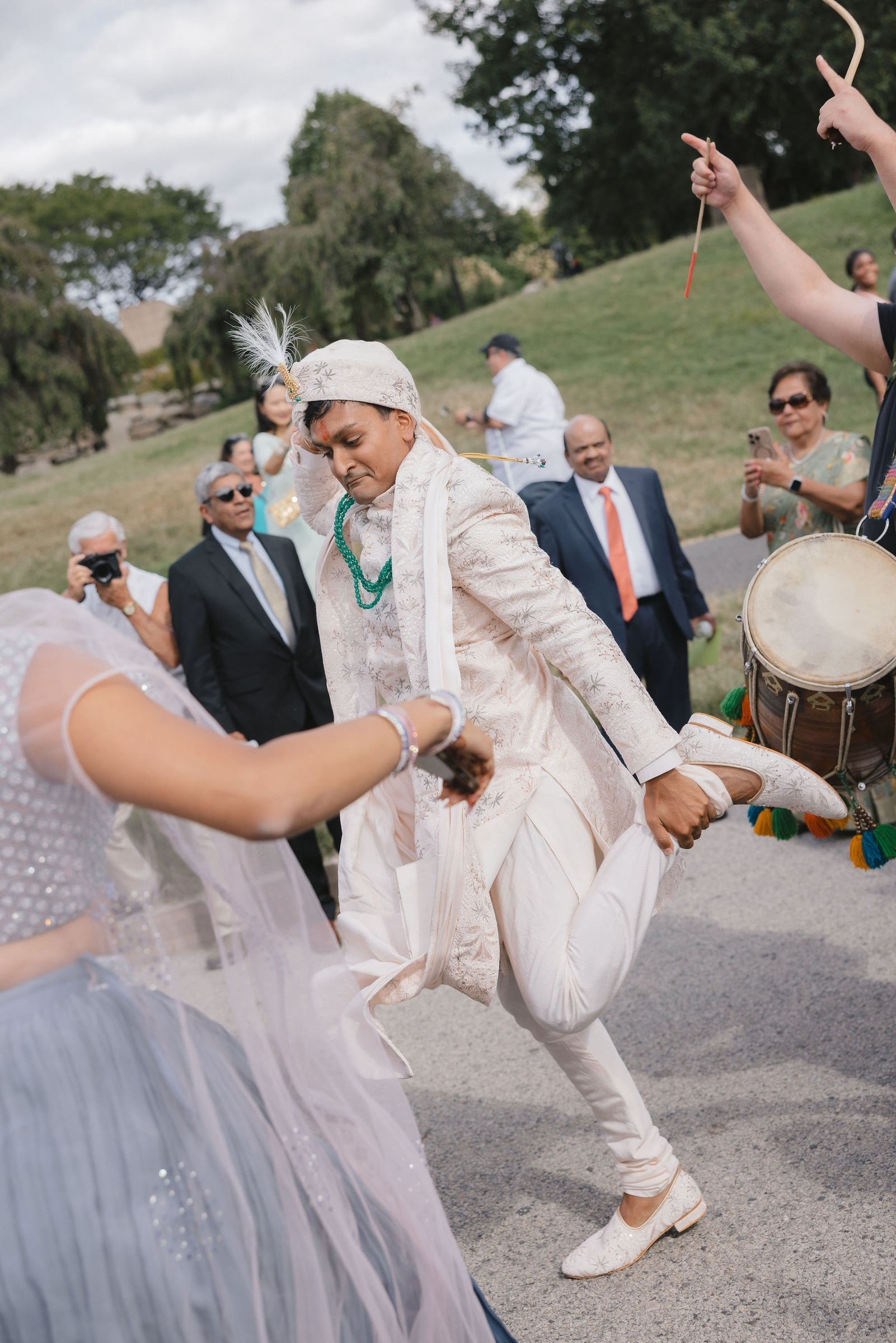 A man dressed in traditional Indian wedding attire kneeling on one knee, holding a woman's hand for a dance. The man is wearing a cream-colored sherwani with matching shoes and a turban decorated with feathers. The woman is wearing a white dress with