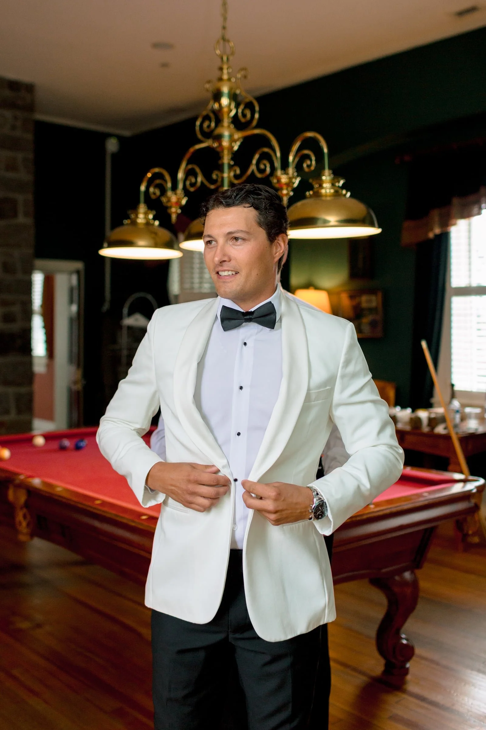 A man dressed in a white tuxedo with a black bow tie stands in a room with a pool table, golden hanging lamps, and a dark green wall.
