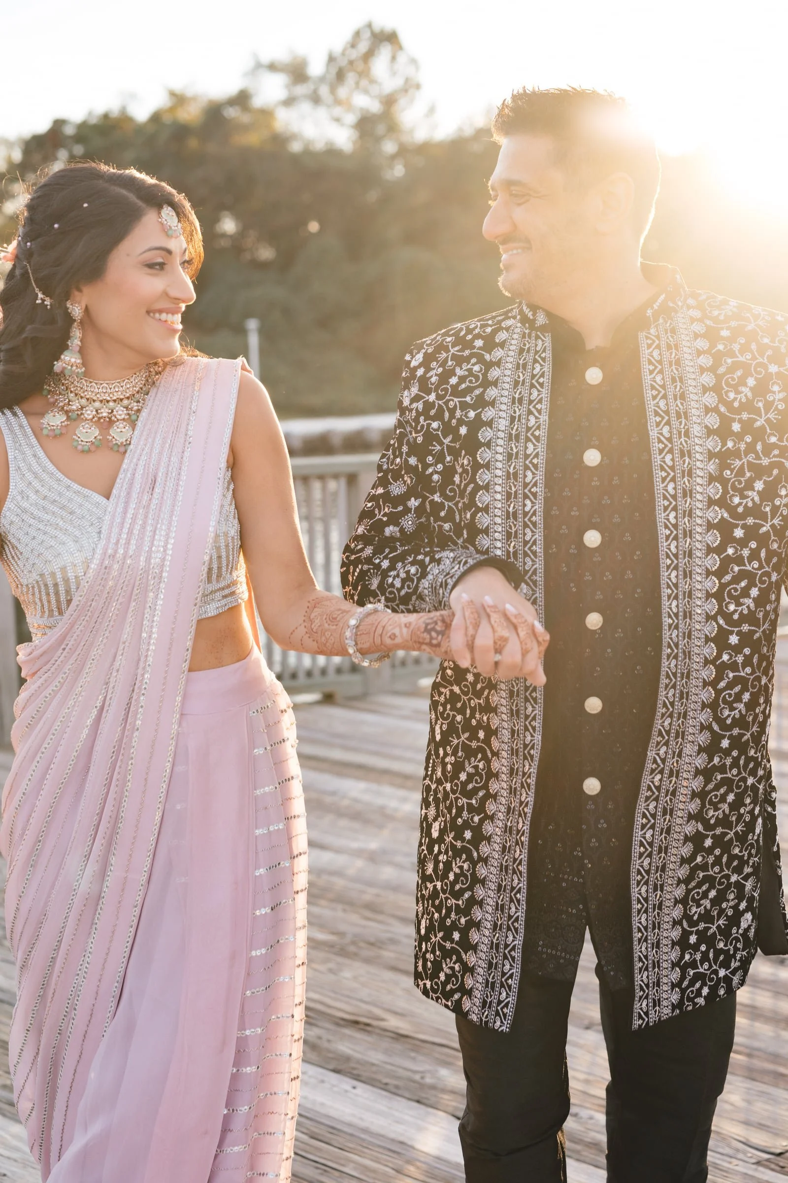 A couple dressed in traditional Indian attire smiles and holds hands on a wooden bridge at sunset, with trees in the background.