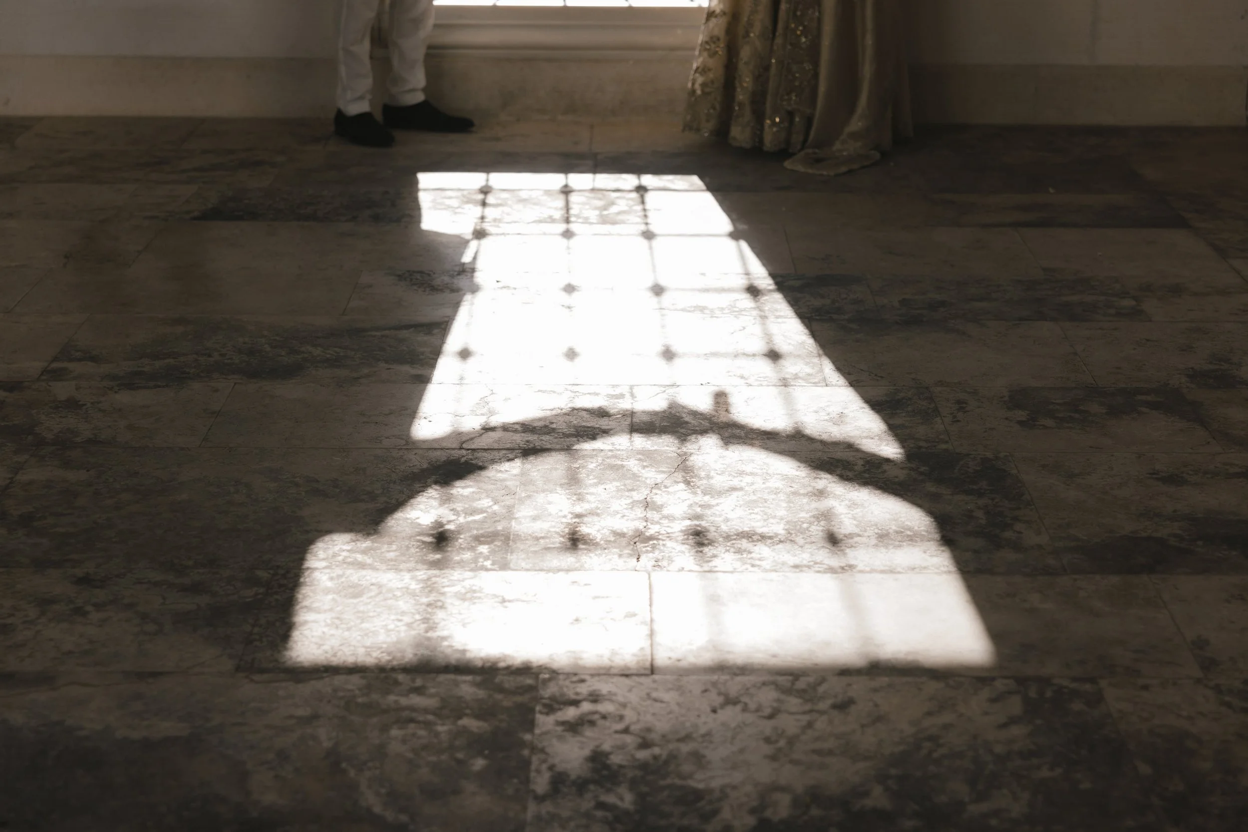 Sunlight shines through a window, creating a grid pattern on a marble floor. Two people are partially visible at the top of the image, one wearing white pants and black shoes, the other in gold and black patterned pants.